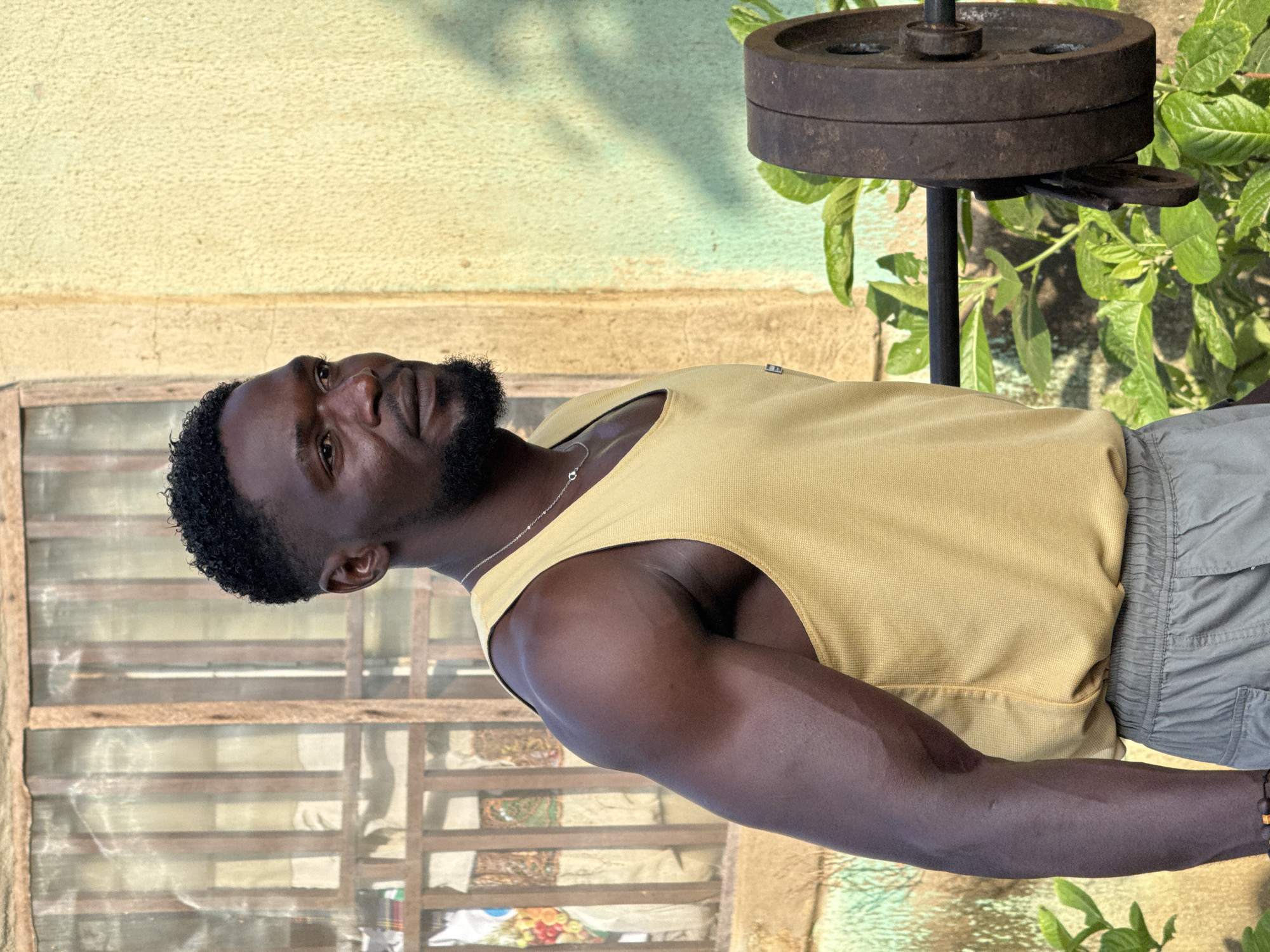 The media file shows a man posing for a photo. He is dark-skinned, with short black hair and a beard. He is wearing a yellow tank top and grey shorts. The man appears to be outdoors, with a textured yellow wall and a window with wooden frames behind him. A weight plate is visible in the upper right corner of the image, suggesting a fitness or weightlifting context. Lush green foliage is also present in the background. The lighting suggests it could be daytime, possibly late afternoon, given the shadows. The man's expression is neutral. There is no visible text in the image. The scene appears to be in Akwanga, Nigeria.
