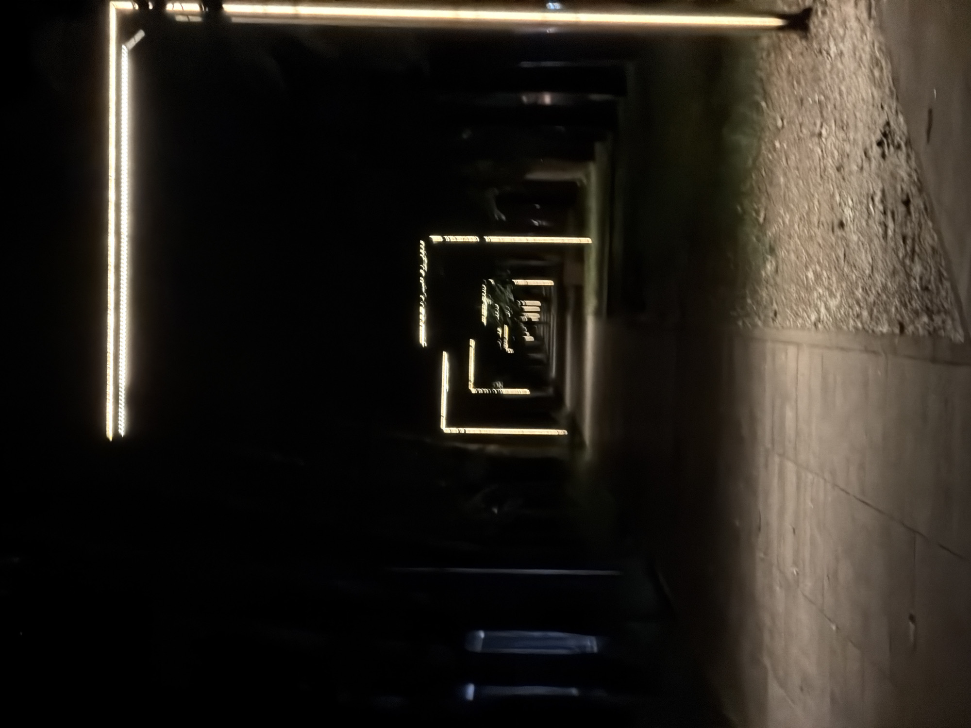 The image depicts a dark, outdoor walkway at night. The walkway is illuminated by a series of rectangular lights that form a tunnel-like effect, receding into the distance. These lights appear to be LED strips, outlining the geometric shapes. To the right of the walkway, there is a rough, textured surface, possibly gravel or a similar aggregate, bordering a paved path. The overall scene is dimly lit, with large portions of the image rendered in deep shadow, suggesting a time after sunset. The location is indicated to be Moscow, Russia, though no specific landmarks are immediately identifiable. There are no people or animals visible in the frame. The atmosphere is one of quiet and possibly solitude, created by the darkness and the repeating pattern of lights. The perspective draws the viewer's eye down the illuminated path, creating a sense of depth.