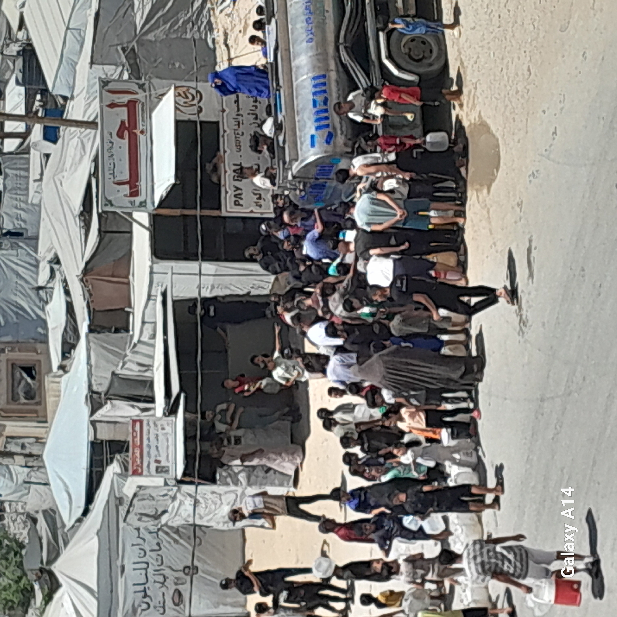 The image depicts a crowded scene in Khan Yunis, Palestinian Territories, likely a displacement camp or an area with temporary shelters. In the foreground, a person in a striped shirt and white shorts walks away from the camera, carrying a red and white cooler. The middle ground is filled with a large group of people, including men, women, and children, gathered around a large tanker truck with Arabic writing and the word "ZAMZAM" in blue letters. Some individuals appear to be receiving water, while others are simply standing or observing. The environment consists of numerous tents and makeshift structures, suggesting a temporary living situation. The ground is dusty and uneven. In the background, more tents and structures extend into the distance. A sign with Arabic text is visible on a building to the left of the tanker truck. The overall impression is one of a communal gathering, possibly for water distribution in a densely populated, resource-scarce area.