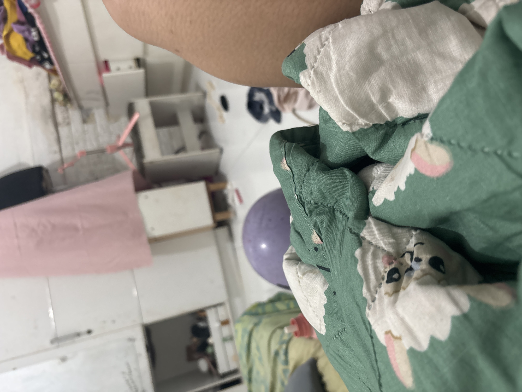 A low-angle, close-up shot captures a section of a person's right thigh and knee, along with a green and white patterned blanket in the foreground. The blanket features a repetitive design of cartoon animal faces, possibly rabbits, with pink accents on their ears. A portion of a purple spherical object, potentially a ball or balloon, is visible behind the blanket. In the background, the scene appears to be an indoor environment, likely a bedroom or storage area. White cabinets with metal handles line a wall, and a pink garment, possibly a dress or skirt, hangs on the left side of the frame. A small white shelf unit with a pink object, possibly a tripod or stand, is situated in the middle ground. The floor is white and scattered with various items, including dark-colored shoes or bags. The overall impression is one of a casually arranged domestic space.