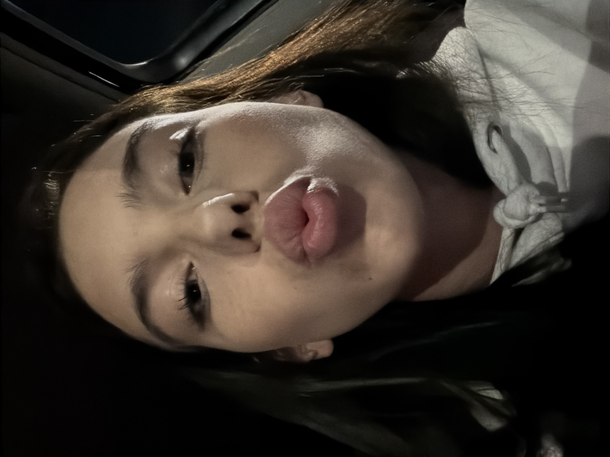 A young woman with long, dark brown hair is captured in a close-up, selfie-style photograph. She is wearing a light-colored hooded sweatshirt with drawstrings. Her lips are pursed in a kissing gesture, and her eyes are open and looking towards the camera. The background is dark, suggesting an indoor or nighttime setting, possibly within a vehicle, given the presence of a dark, curved edge on the right side of the frame that resembles part of a car interior. The lighting appears to be artificial, casting shadows and highlighting the contours of her face, particularly her cheekbones and lips. There is no discernible location in Sofia, Bulgaria, evident from the visual information provided.