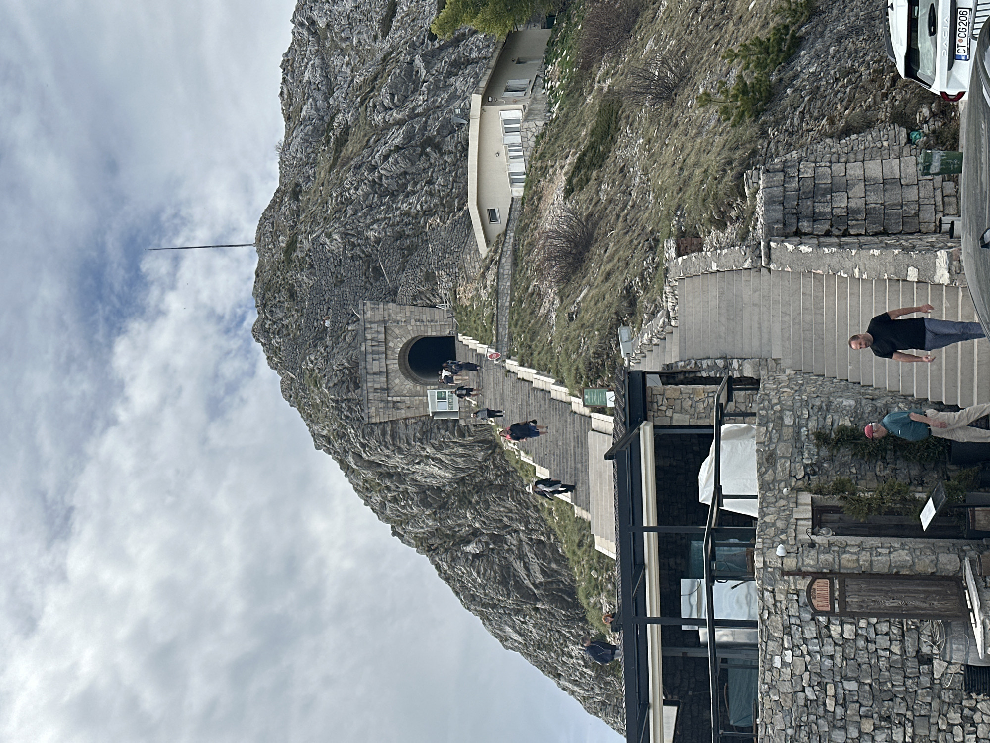 The image shows the entrance to a tunnel in the mountain, leading up to the Mausoleum of Petar II Petrovic-Njegos in Lovćen National Park, near Mali Bostur, Montenegro. A long set of stone stairs ascends from the base towards the tunnel entrance, with several people walking up or down. A building with a modern glass-and-stone structure stands on the right side of the staircase. Two cars are parked on the right. The sky is cloudy. A thin metal rod rises from the top of the mountain above the tunnel.