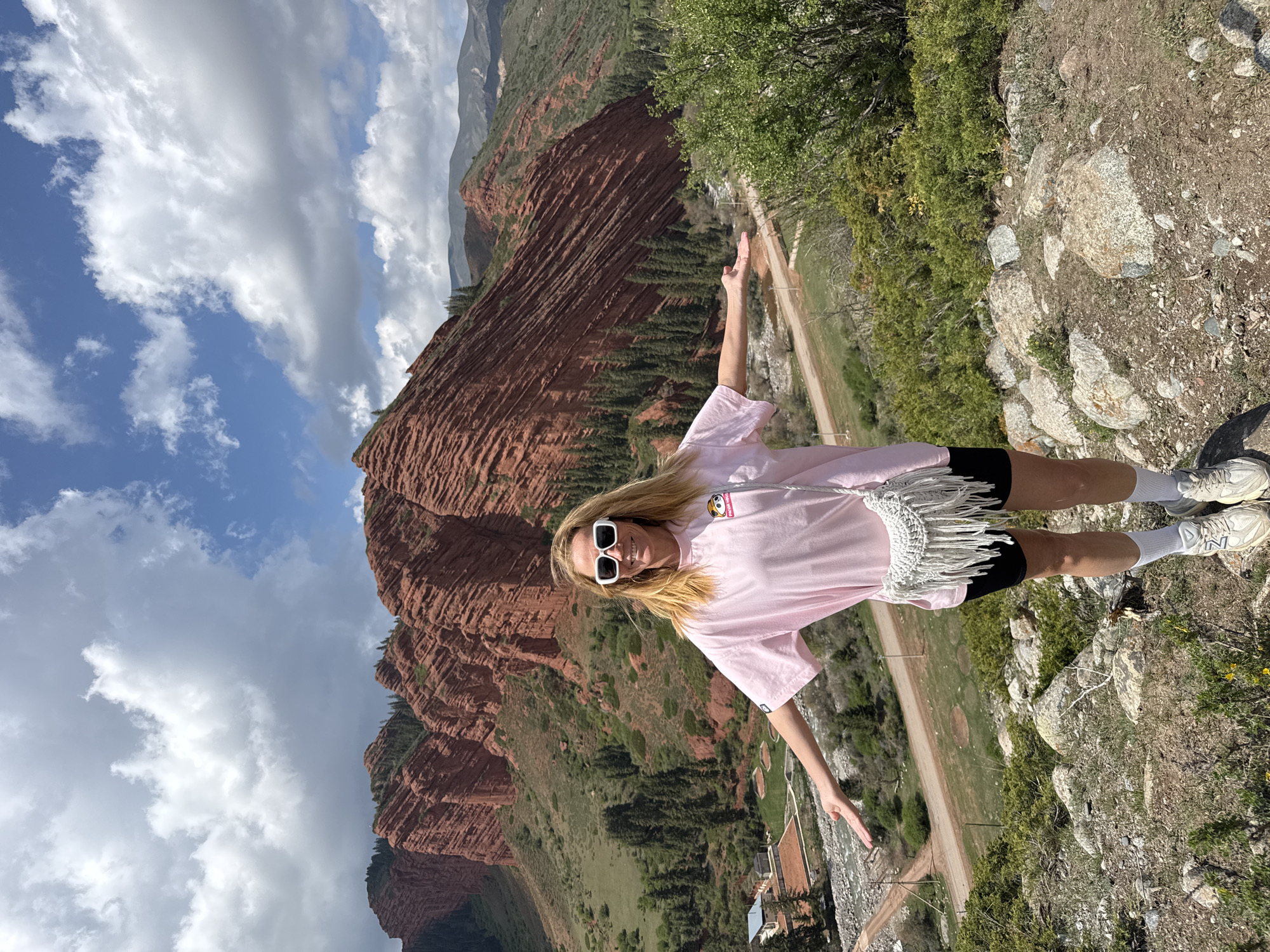 A blonde woman, appearing in her 20s or 30s, stands posing outdoors in a striking mountainous landscape. She is wearing a light pink oversized t-shirt, black shorts, white athletic socks, and light-colored New Balance athletic shoes. White rectangular sunglasses are on her face, and a light-colored, possibly white, crocheted or woven bag with fringe is worn across her body. Her arms are slightly outstretched, and she looks directly at the camera with a gentle smile. A small cartoon-like patch is visible on her pink t-shirt.

The background features prominent, rich red, stratified rock formations that form dramatic cliffs and mountains, characteristic of the Dzhety-Oguz area in Kyrgyzstan. Portions of these mountains are covered in vibrant green foliage, including trees and bushes. Below the cliffs, a dirt road winds through the valley, and a few small structures are discernible in the distance. The foreground consists of rocky, earthy ground with scattered green vegetation.

The sky above is partly cloudy, with patches of clear blue contrasting with bright white clouds, indicating it is daytime, likely late morning or afternoon. The overall scene conveys a sense of adventure and appreciation for the natural beauty of the location. The "New Balance" logo is visible on the side of her right shoe.