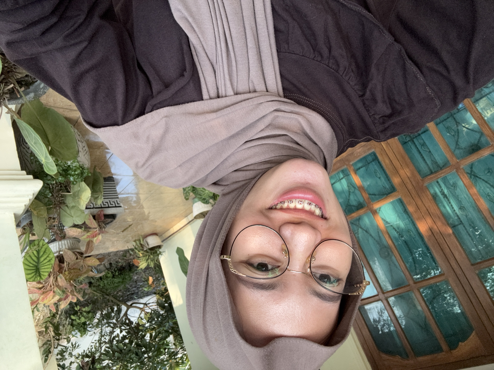 A young woman with light skin and brown eyes is pictured in a selfie taken outdoors, likely in the city of Gadingrejo, Indonesia. She is wearing a brown hijab and a dark brown long-sleeved shirt. She has on round, gold-rimmed glasses and braces on her teeth, which are visible as she smiles. Behind her, a wooden door with glass panes is visible on the left side of the frame. The right side of the image shows a garden with various potted plants and a white pillar. The ground appears to be tiled. The overall lighting suggests it is daytime.