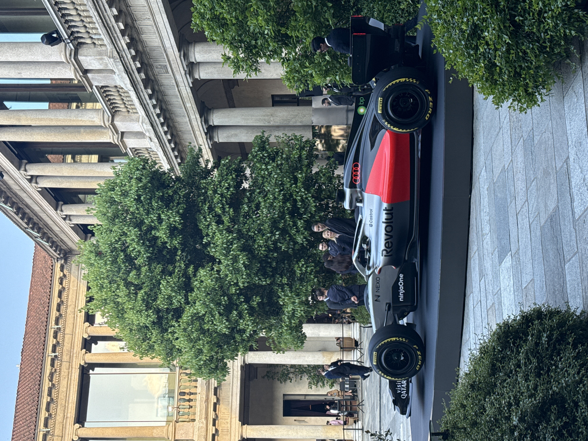 An outdoor scene in Milan, Italy, captures a Formula 1 race car on display during the daytime. The car, painted in silver-grey, red, and black, sits on a dark platform, featuring prominent branding such as "Revolut," "ninjaOne," "N NEXO," "Castrol," "bp," "AUDI," and "visit QATAR." Its tires are clearly marked "IRELLI" and "P ZERO."
The setting is an elegant urban environment, characterized by a grand building with classical architecture, including large stone columns, ornate cornices, and detailed window frames. Lush green trees and bushes are interspersed throughout the area, providing a natural contrast to the built environment. In the background, hints of outdoor café seating suggest a public square or boulevard.
Several people, mostly dressed in formal or business attire, are visible around the car, observing it or conversing, indicating a public event or promotional activity. The bright, natural light suggests a clear day, possibly late morning or afternoon. The overall atmosphere appears calm and appreciative, focusing on the impressive display of the F1 car against the backdrop of historic Italian architecture.