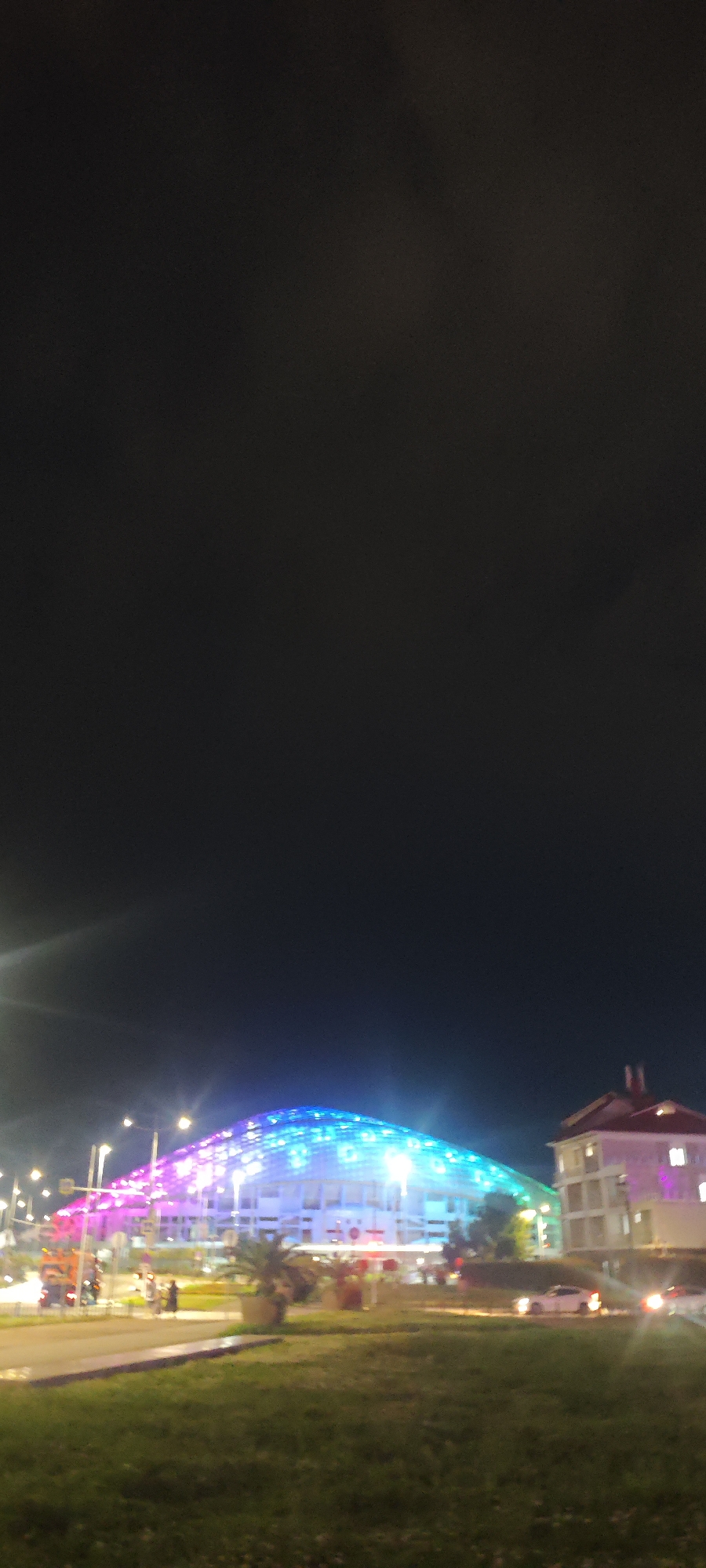 At night in Sirius, Russia, a colorful illuminated dome structure is visible behind a grassy lawn. A two-story building sits adjacent to the dome on the right. Streetlights illuminate the road and buildings, and a few people are visible on the sidewalk. Cars are driving on the road.