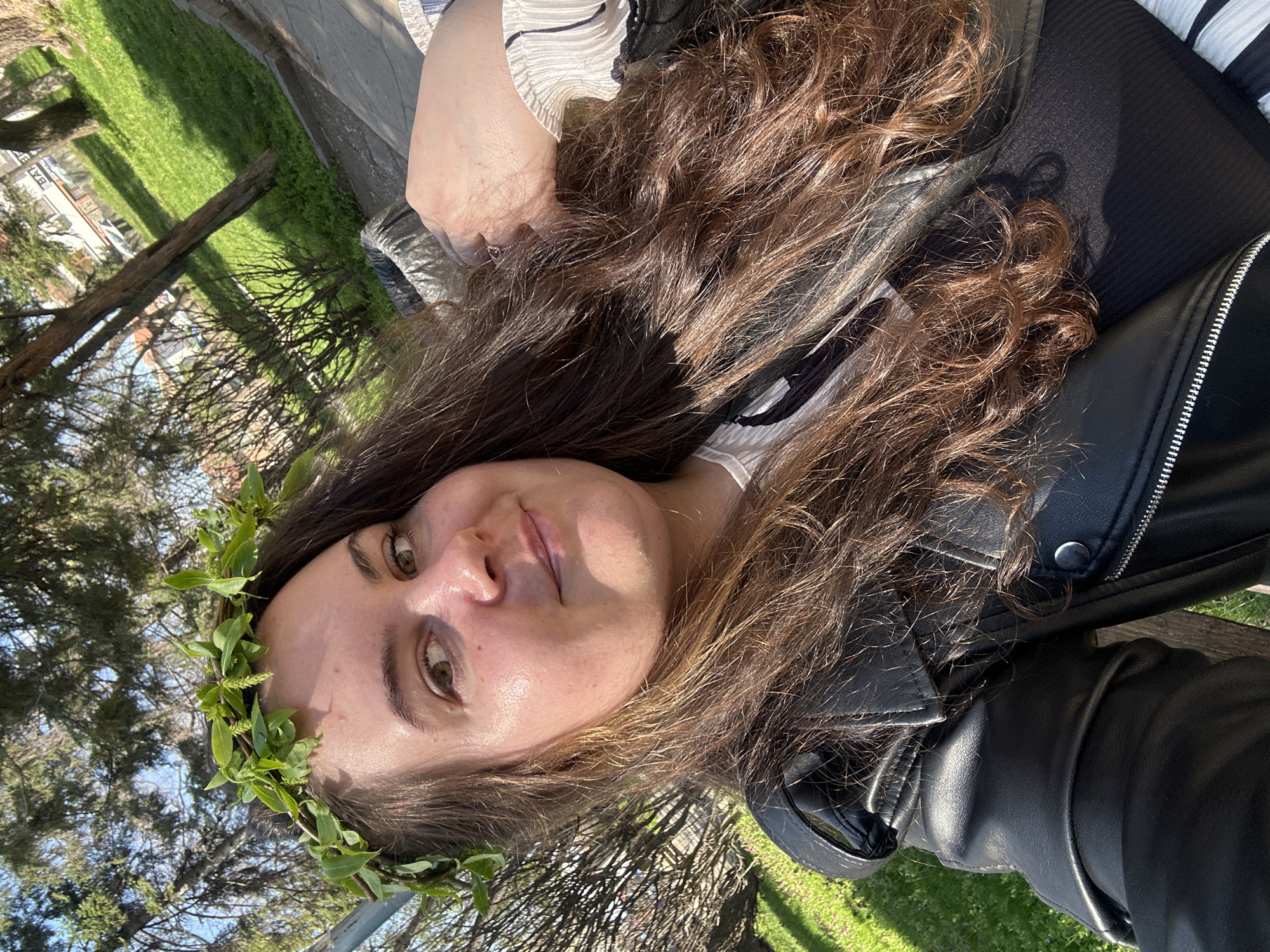 A selfie taken by a light-skinned woman with long, curly brown hair wearing a crown of green leaves on her head, a striped shirt, and a black leather jacket. The background shows trees, grass, and sky, suggesting an outdoor setting, possibly a park. It is not possible to determine the location of the scene as Plovdiv with the provided information.