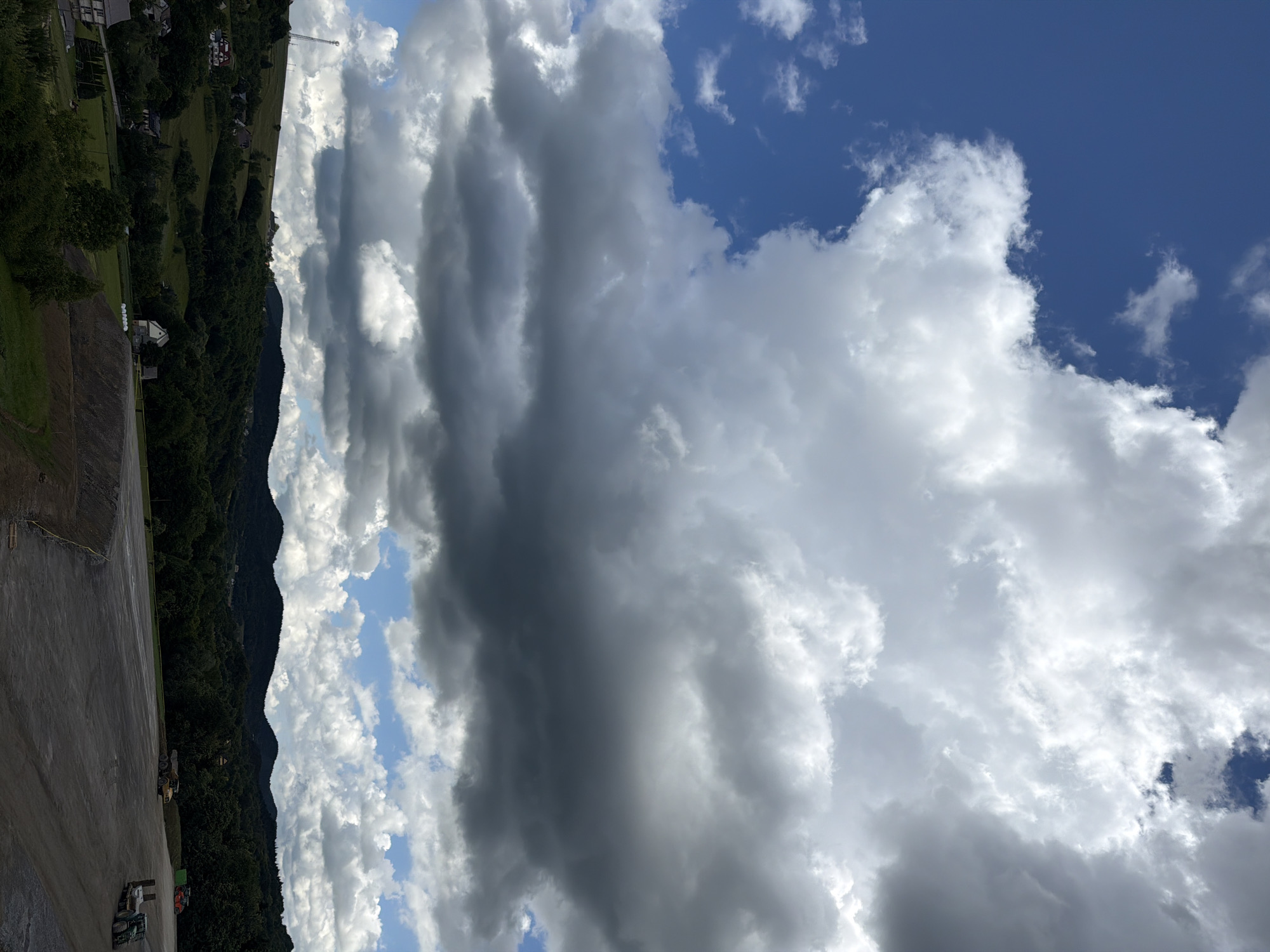 The image depicts a view of the city of Stróża, Poland, on a day with dramatic cloud formations. The foreground is dominated by a large, paved area, possibly a construction site or a parking lot, with some heavy machinery visible. To the left and in the background, rolling green hills covered in dense forest rise up. Scattered amongst the trees are houses and buildings, indicative of a rural or semi-rural setting. The sky above is a dynamic mix of bright blue patches and large, imposing clouds, some of which are dark and heavy, suggesting an impending storm or recent rainfall. The overall impression is one of a vast landscape under a changeable sky, with human activity present in the form of machinery and scattered structures.