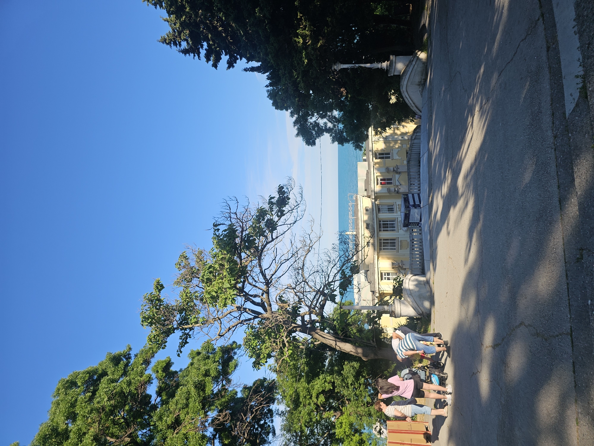 The image shows a group of four people, including two adults and two children, gathered near a baby stroller on a paved pathway. They are surrounded by lush green trees and a clear blue sky. In the background, a light yellow building with white window frames and a fence can be seen, with the ocean visible beyond it. The building appears to be a historical or architectural structure, and the overall scene suggests a pleasant, sunny day in a scenic location, possibly a park or a waterfront promenade. One of the adults is standing next to the stroller, while the other is looking down at it. One child is standing beside the stroller, and another is interacting with the stroller. The trees in the foreground are large and mature, with dense foliage, and cast shadows on the pathway. The ocean in the background is calm and blue, with a faint horizon line visible. The light yellow building in the background is the focal point of the architecture.