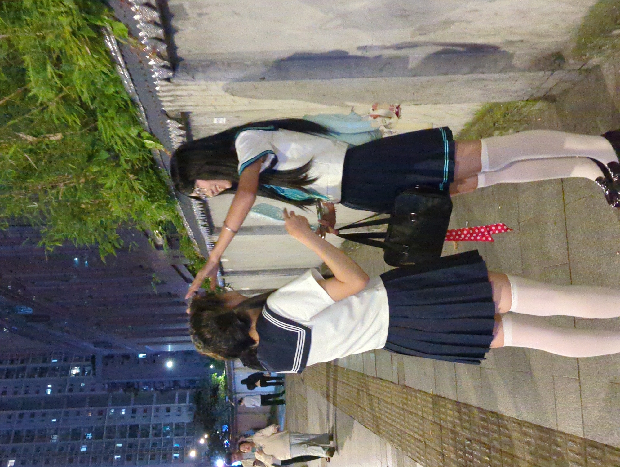 Two women in school uniforms stand outside in Liangjiang, China at night, in front of a large apartment complex. The woman in the foreground wears a white shirt with a dark navy blue sailor collar, a navy blue pleated skirt, and white knee socks. The woman in the background wears a white shirt with blue trim on the sleeves, a navy blue pleated skirt with blue trim, and white knee socks. She touches the hair of the woman in the foreground. A black satchel lies on the ground between the two women. Additional people stand in the background.