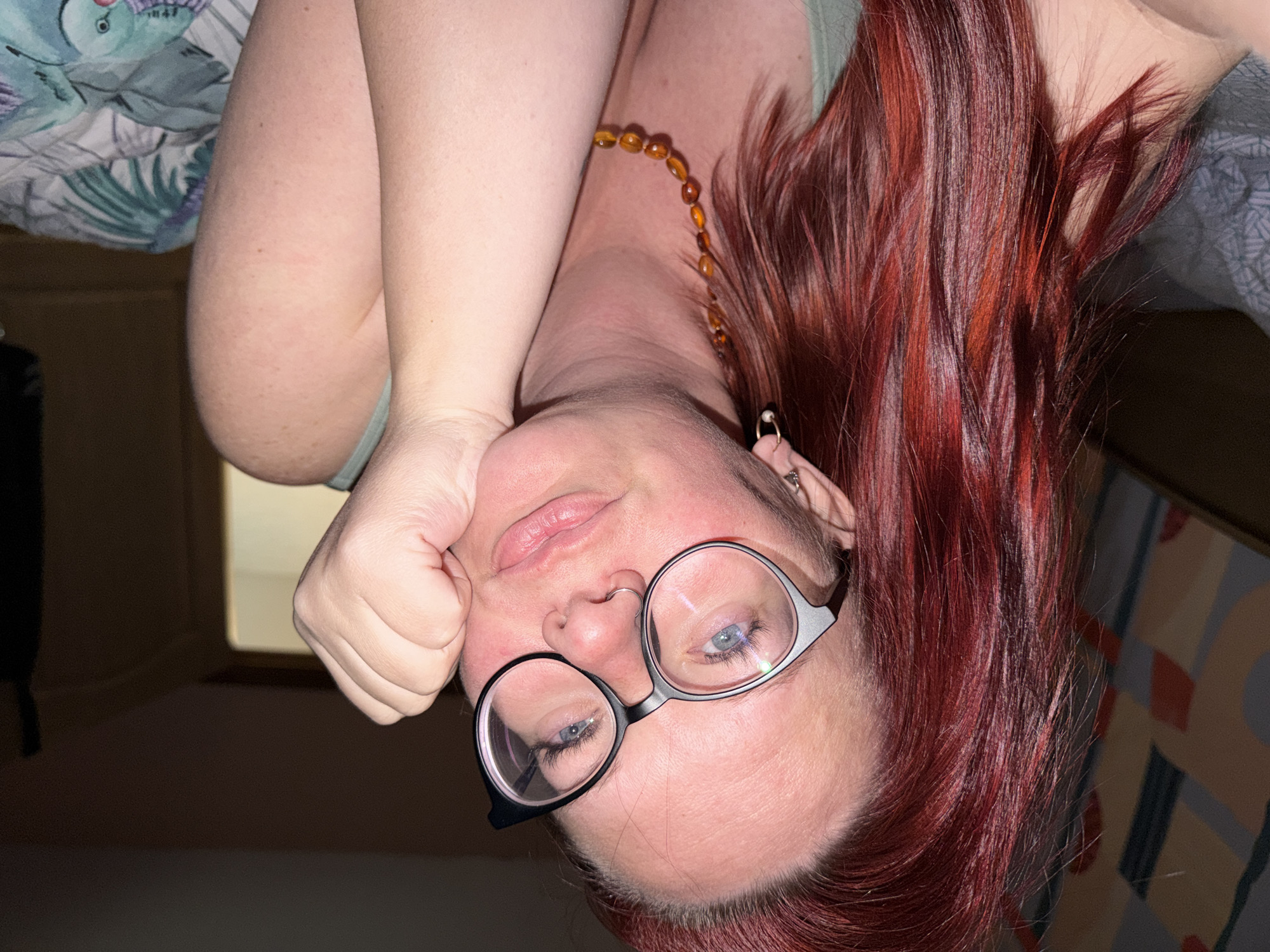 A Caucasian woman with red hair and round glasses is upside down, looking at the camera. She has blue eyes, a nose ring, and pierced ears. She is wearing a necklace with brown beads. Her right hand is balled into a fist and resting on her forehead. The background includes a bed with floral patterned sheets and a patterned wall. The location is not discernible.