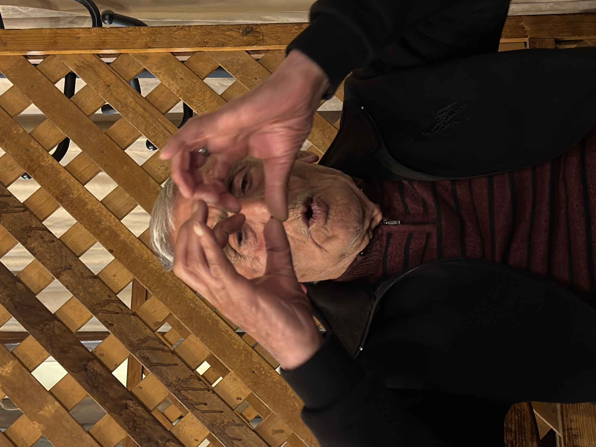 A photo shows a senior man indoors, with his hands making a heart shape over his face. He has gray hair and wrinkled skin. He's wearing a black jacket over a maroon shirt with black stripes. Behind him is a wooden lattice. The image is taken at an odd angle. The location is presumed to be Yerevan, Armenia.