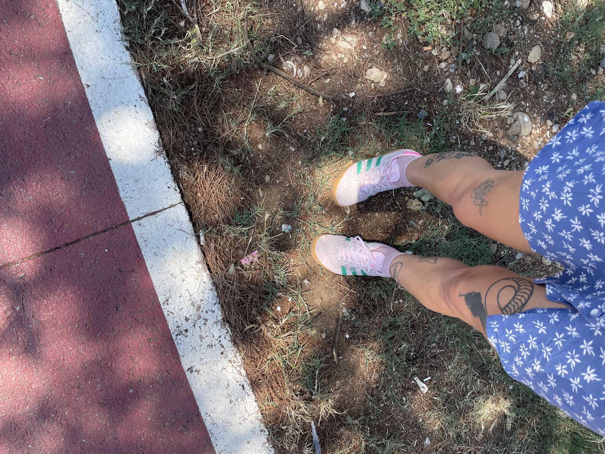The image shows a person's legs from a top-down perspective, standing on a patch of dry grass and pine needles adjacent to a red surface with a white border. The person is wearing pink Adidas sneakers with green stripes and light blue shorts with a white floral pattern. There are tattoos on the person's legs, including what appears to be a snake on the left thigh and another on the right calf. The environment suggests an outdoor location, possibly a park or recreational area in Podgorica, Montenegro, given the presence of dry vegetation and a paved surface with a border, common in such spaces. The lighting indicates it is daytime, with shadows cast by overhead foliage. There are no other people visible in the frame.