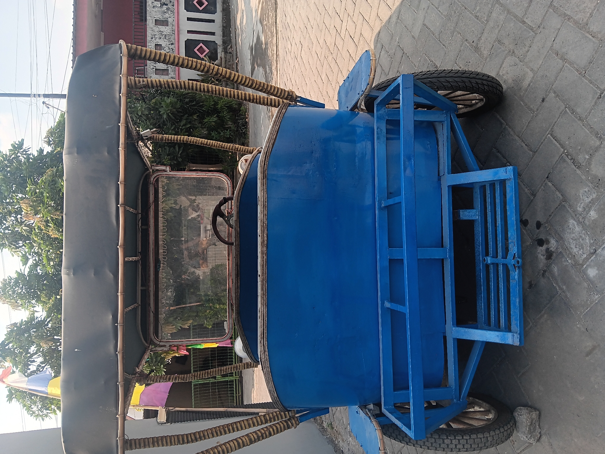 A blue, open-air, three-wheeled vehicle, resembling a modified rickshaw, is parked on a paved walkway in Tlogo, Indonesia. The vehicle features a black canvas roof supported by bamboo poles, a wooden body painted blue, a metal frame with footrests, and two large rear wheels. A steering wheel is visible through the windshield. In the background, trees, a fence, and a residential building with patterned walls are visible. A flag with red, white, blue, and yellow sections is flying above the vehicle. The lighting suggests it is daytime.