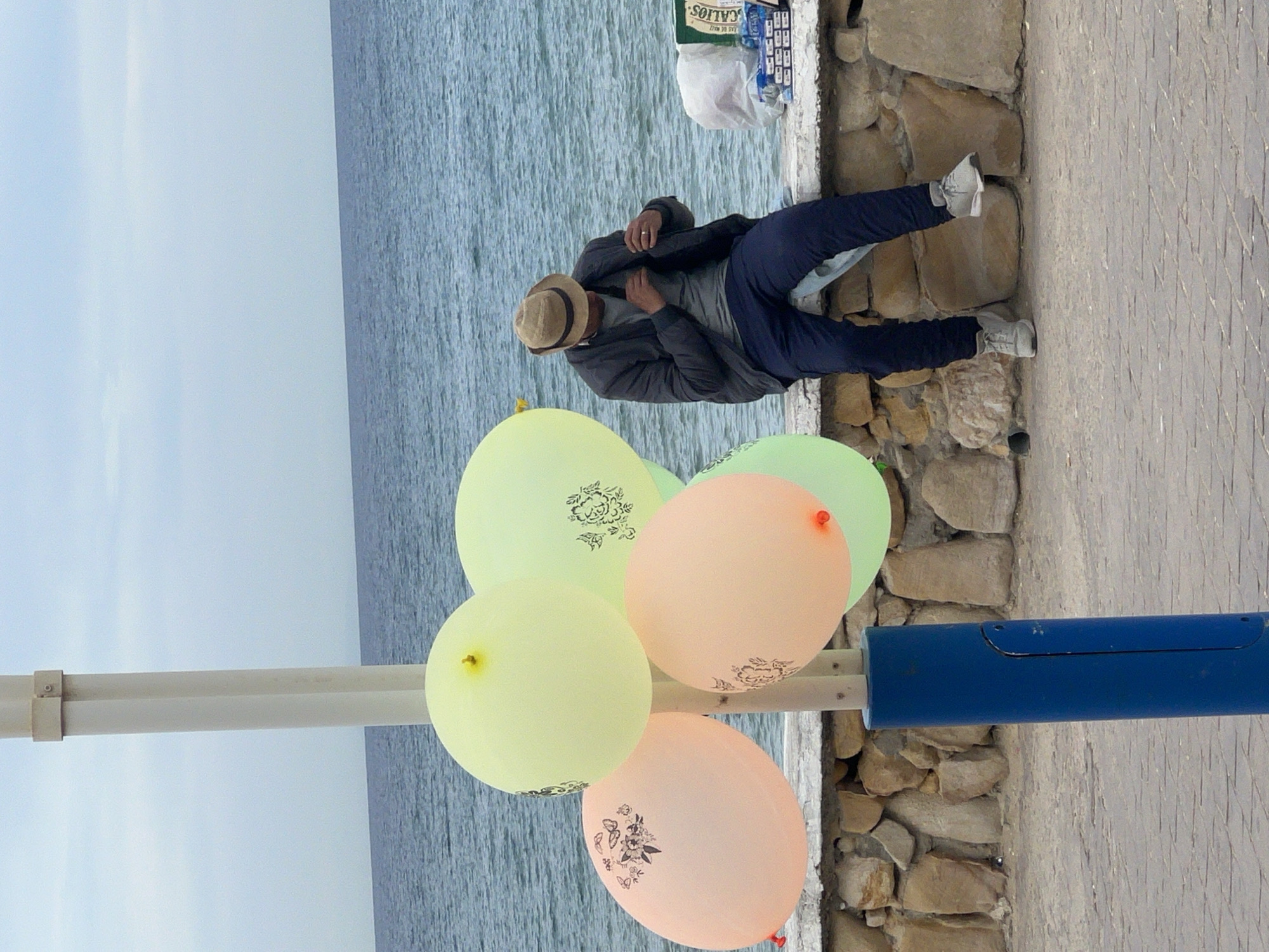 A man with a fedora and blue pants sits on a stone wall overlooking the ocean. Beside him are several helium balloons attached to a metal post. The location is likely in M'Diq, Morocco, given the context.