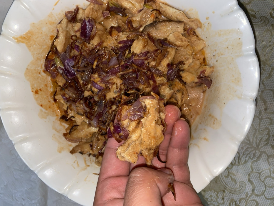A close-up shot displays a white plate filled with a mixture of cooked food. The primary components appear to be shredded or diced chicken, generously topped with sautéed red onions. The plate has some staining, suggesting the food has been saucy. A human hand is holding a portion of the chicken and onions, lifting it slightly from the plate. The person's fingers appear slick with grease or sauce.