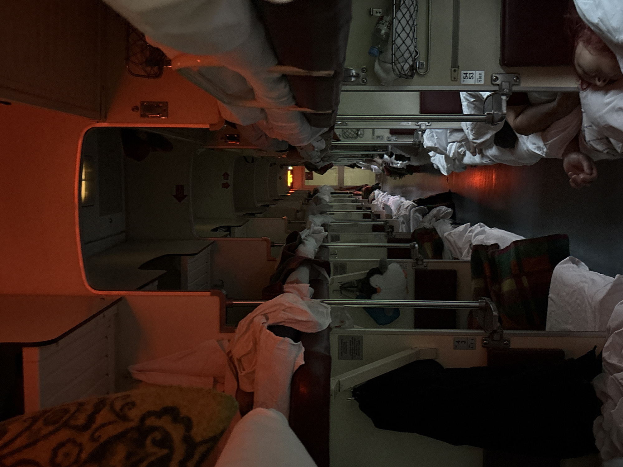 The image depicts the interior of a train car, likely a sleeper compartment, illuminated by dim, reddish lighting. The car is lined with multiple bunk beds on both sides, most of which appear to be occupied or prepared for occupancy with white linens. A corridor runs down the center of the car, reflecting the orange light. In the foreground, a person is sleeping on a lower bunk, with their head resting on a pillow and their body partially covered by bedding. Other passengers are visible in the distance, lying on their bunks or walking in the corridor. Various personal items, such as a black jacket hanging on a hook and a stuffed penguin, are also present. The train car appears to be in transit, with the environment suggesting a long-distance journey. The presence of bunk beds and the overall layout are characteristic of a Russian train car. The specific location in the city of Korzhava, Russia, cannot be determined from the visual information alone.