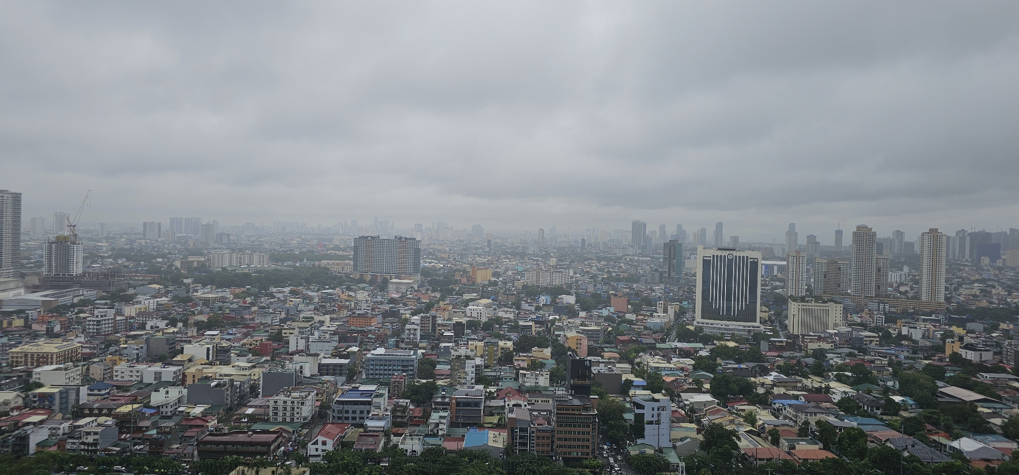 The image is a panoramic, high-angle view of the city of Makati, Philippines, on a heavily overcast day. The sky is filled with dense, gray clouds, obscuring any clear sunlight and giving the scene a subdued, muted color palette. The cityscape is dominated by a dense collection of buildings, ranging from low-rise residential structures with colorful rooftops to mid-rise and numerous high-rise commercial and residential towers.
In the foreground, a cluster of smaller, densely packed buildings with varied roof colors are visible. Moving towards the mid-ground and background, the density of buildings increases, and the structures become taller. Prominent among these are several modern skyscrapers, characterized by their sleek glass facades and distinct architectural designs. One notable building in the mid-right is a tall, rectangular skyscraper with a dark facade and vertical white stripes. To its left, a cluster of three very tall, light-colored buildings stands out, appearing to be residential condominiums. Further into the distance, a hazy urban sprawl extends, with countless buildings fading into the atmospheric conditions.
Construction cranes are visible in several locations, indicating ongoing development and urban expansion within the city. The overall impression is one of a bustling, densely populated metropolis with continuous growth. There are no discernible individuals or specific human activities visible in the frame due to the distance and the image's focus on the urban landscape. The environment appears to be a typical urban setting, characterized by concrete structures, roads, and scattered pockets of greenery, likely trees and vegetation interspersed within the urban fabric. The weather appears to be calm, with no immediate signs of rain, but the heavy cloud cover suggests the possibility of precipitation.