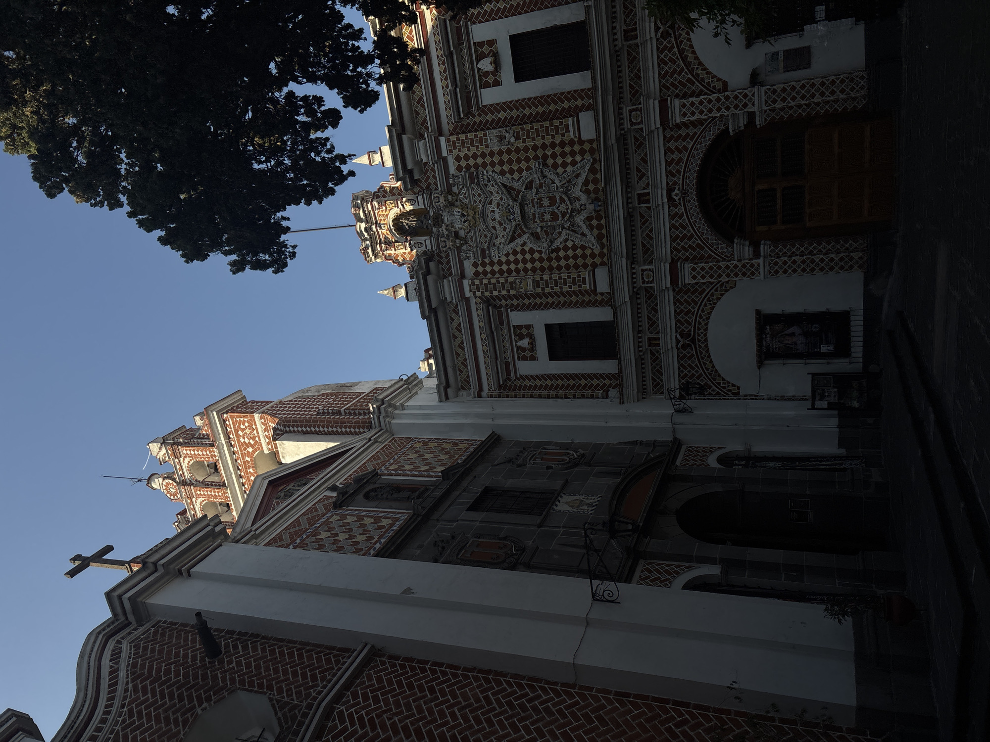 The image shows a low-angle view of a church facade in Puebla, Mexico, identified by its characteristic red brick and white decorative patterns. The sky is visible above, with a dark tree obscuring part of the upper left corner. The building features multiple levels and windows, with an ornate central section decorated with architectural embellishments. A cross is positioned atop a section of the building. The facade appears partly shadowed, suggesting the time of day or angle of the sun. No people are visible in the frame. The overall scene suggests a historical or religious site in the urban landscape of Puebla.