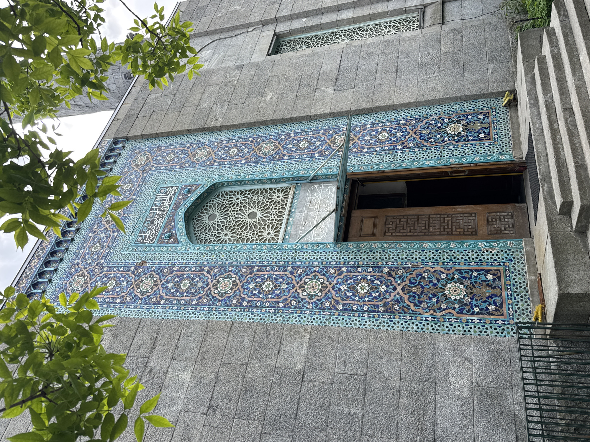 The image displays a detailed view of a building facade, showcasing an impressive section of ornate ceramic tile work set against grey stone blocks. The tile work, predominantly in shades of blue, turquoise, white, and dark blue, features intricate geometric patterns and stylized floral motifs, characteristic of Islamic art.
Within this tiled area, an arched window is covered by a delicate, white screen with a complex star-shaped geometric design. Adjacent to it, an open window or doorway reveals a dark interior and some dark wooden elements with carved patterns. Above this main tiled section, a smaller rectangular window is also fitted with a similar geometric screen. Arabic calligraphy is integrated into the tile design, especially around the arched window.
The surrounding parts of the facade are constructed from large, rough-textured grey stone blocks. To the far right, a prominent concrete or stone structure with a distinct stepped, ridged texture is visible. Bright green leaves from a tree are positioned in the foreground on the left and top left, partially obscuring the building, suggesting an outdoor setting with vegetation. A dark metal railing is visible at the bottom right.
The scene depicts the exterior of an architecturally significant building, likely a mosque given the distinct tile work and design, under an overcast or bright cloudy sky during daytime. No specific activities or people are visible. The location context indicates this building is in Saint Petersburg, Russia.