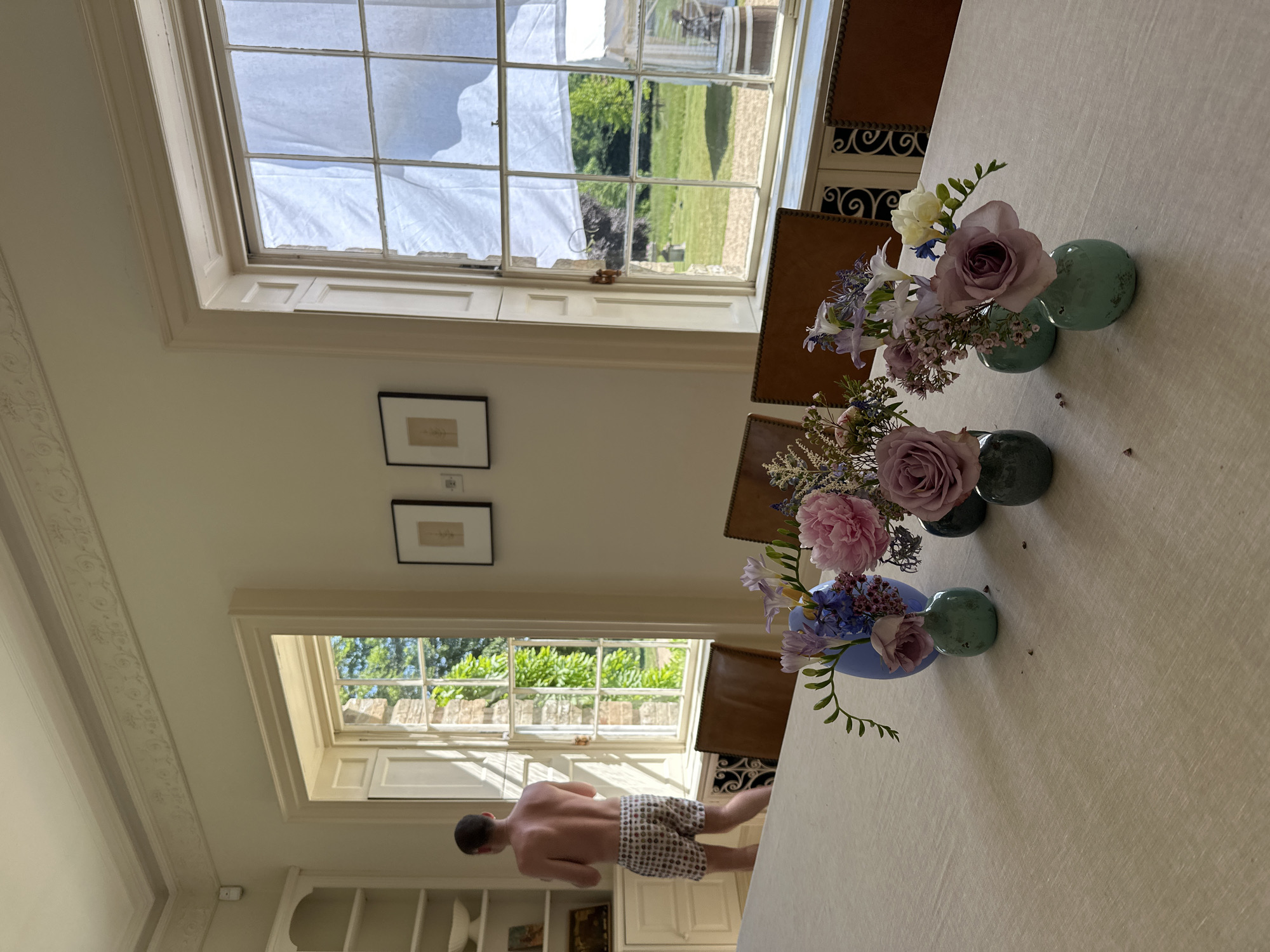 A slightly tilted interior shot features a room with two windows, a table adorned with floral arrangements, and a partially visible person in the lower section. The windows offer views of exterior scenes, one obscured by a white covering. Two framed artworks hang on the wall between the windows. Three floral arrangements are centered along the table, each consisting of various flowers in small vases. In the lower part of the frame, a person with a bare torso and patterned shorts is walking away from the camera near the bottom window. The environment includes architectural details and built-in shelving. The location appears to be in the city of Unknown, Unknown.
