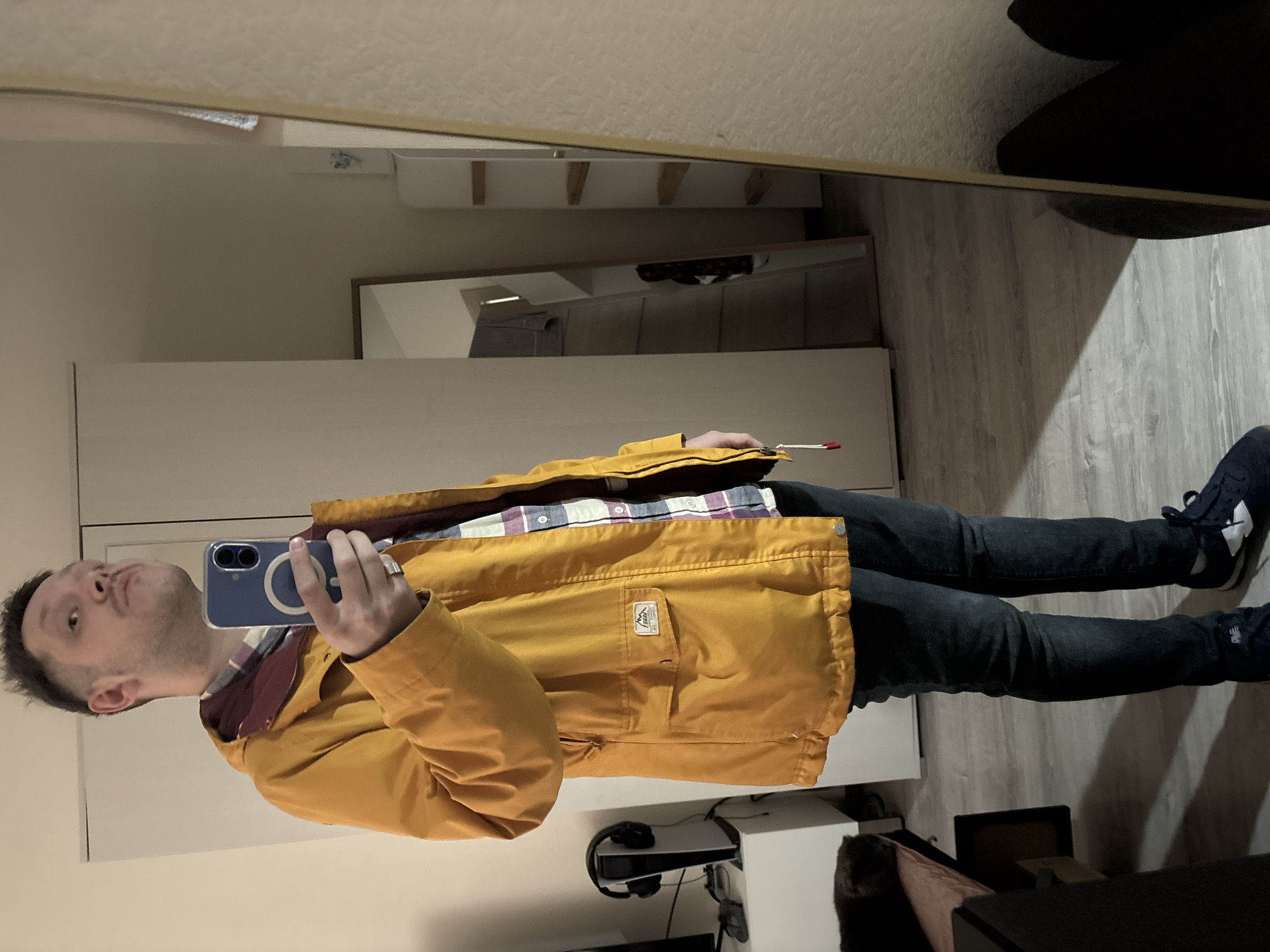 A man is taking a selfie in a mirror. He is wearing a yellow jacket, a plaid shirt, and dark jeans with blue shoes. The background shows a room with a white dresser, a mirror, and what appears to be a chair. The location in Saint Petersburg, Russia, is not verifiable from the image.