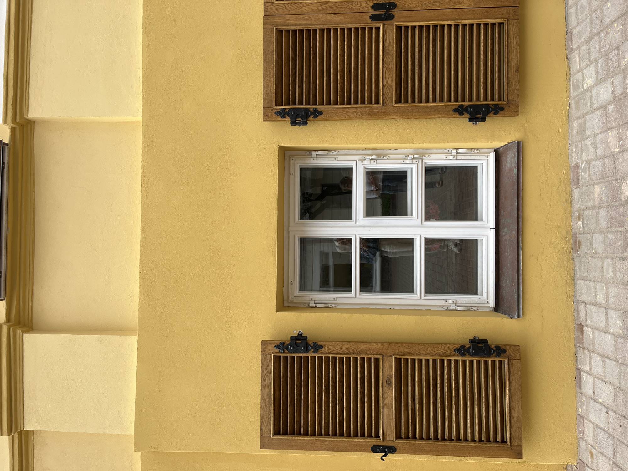 A medium shot captures a yellow-painted building facade featuring a central white-framed window with wooden shutters on either side. The window is divided into six panes, and reflections of two people can be seen within the glass. The shutters are made of wooden slats and secured with black metal hinges and latches. The building's exterior has decorative architectural elements. The foreground shows a cobblestone pavement. The location is a street in the city of Vitsebsk, Belarus.