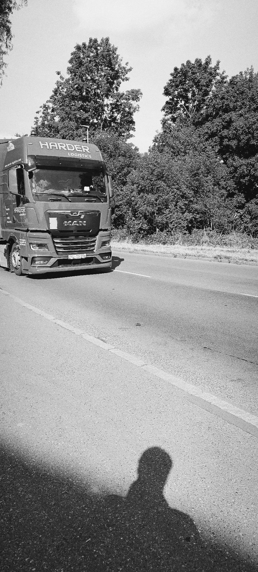 A black and white, eye-level, medium shot shows a MAN semi-truck with "HARDER LOGISTICS" written on its side, driving on a road. The truck is a dark gray color. The truck's front grill is clearly visible, with the "MAN" logo. The license plate reads "NU DS 556." The truck is traveling on an asphalt road with a white line dividing the lanes. Trees and foliage line the right side of the road, and the sky is visible in the background. A shadow of a person holding a camera is cast on the road in the foreground, obscuring the bottom left corner of the image. The scene appears to be captured on a sunny day. The location is identified as Waiblingen, Germany.