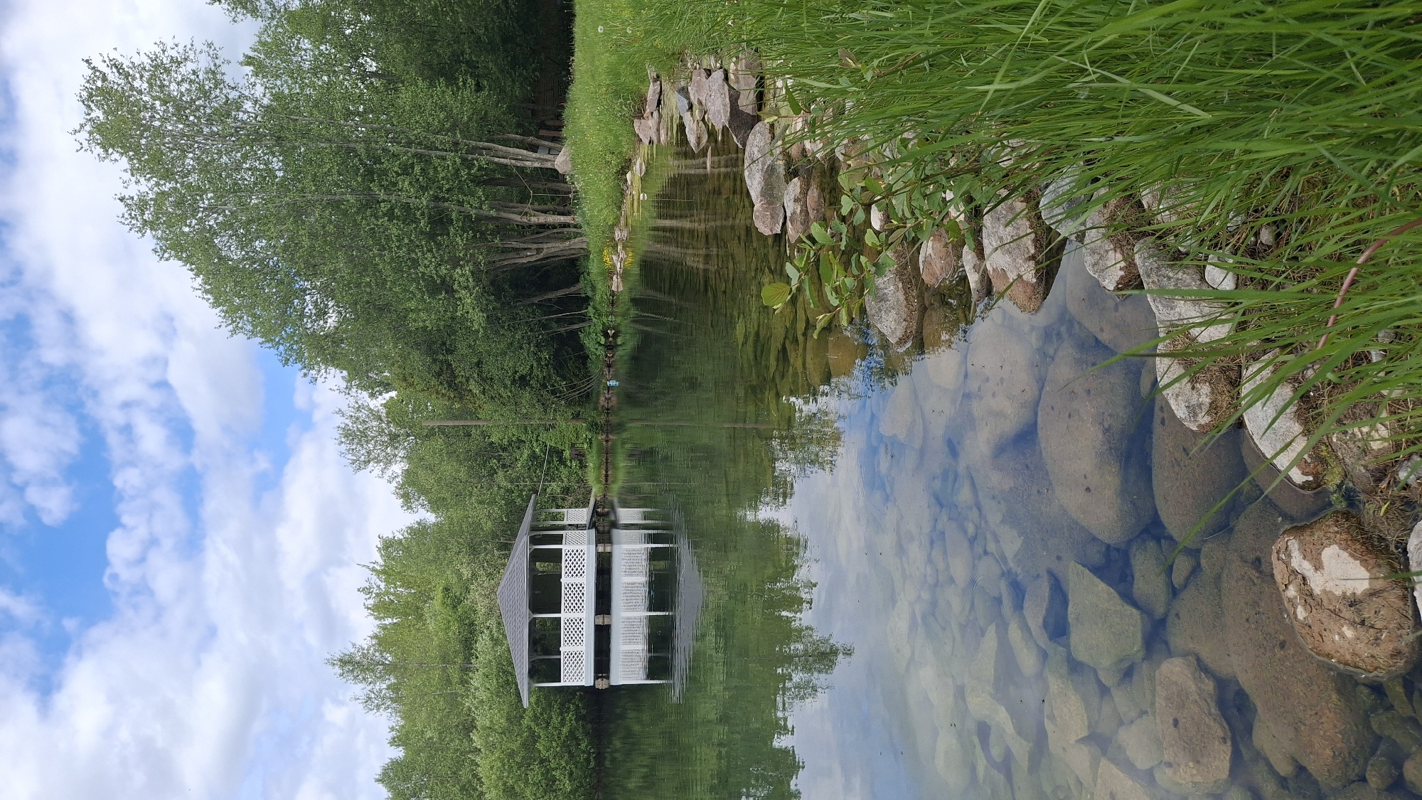 The image captures a serene outdoor landscape in Kipen, Russia, featuring a tranquil body of water, likely a pond or calm river, reflecting its surroundings. The water, clear enough to reveal submerged rocks and pebbles at its bottom, occupies a significant portion of the frame. Its surface provides a near-perfect mirror image of the bright blue sky dotted with white clouds and the dense, lush green trees lining the banks.
Along the immediate foreground and right side of the water, the bank is lined with various sized rocks and boulders, interspersed with vibrant green grass. On the opposite side of the water, a distinctive white gazebo with a dark, possibly gray, roof stands amidst a verdant backdrop of numerous trees. The gazebo's reflection is sharply defined in the still water.
The scene depicts a pleasant, bright daytime, likely late morning or early afternoon, under partly cloudy skies. No people or specific activities are visible, emphasizing the peaceful and undisturbed nature of the environment. The overall impression is one of natural beauty and tranquility in a well-maintained setting. There is no visible text in the image.