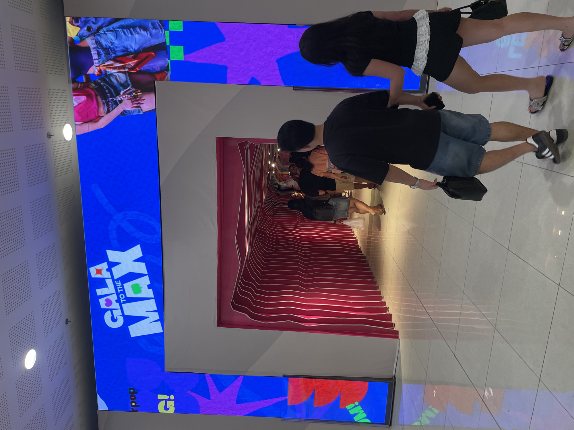 The image depicts an indoor scene within a shopping mall or event venue, identified as SM Lanang Premier in Davao City, Philippines, during an event called "GALA TO THE MAX." A large, bright blue digital billboard spans the upper part of the scene, displaying the text "GALA TO THE MAX" in prominent white lettering with a heart icon replacing the first 'A', a star icon replacing the second 'A', and a green square replacing the 'A' in 'MAX'. The left side of the billboard shows partial text "rpop" and "G!", while the right side displays cropped images of individuals, including a person wearing a pink crop top and denim shorts, and another in blue jeans with an orange and white patterned scarf draped from their waist.
Below the billboard is a distinctive entrance to a curved, tunnel-like pathway. The exterior of this tunnel is a solid pink, and its interior walls feature prominent white, wavy lines against a pink background, creating a visually dynamic effect. Multiple individuals are visible entering and moving through this tunnel, suggesting an event or exhibition space.
In the foreground, a man and a woman are walking away from the viewer towards the tunnel entrance. The man, on the left, wears a dark short-sleeved t-shirt, blue denim shorts, and dark athletic shoes with white stripes. He carries a black handbag in his left hand and appears to be holding a dark object, possibly a phone, in his right. The woman, on the right, wears a black short-sleeved top with a white lace-trimmed peplum, black shorts, and metallic silver slip-on sandals. She carries a black handbag in her left hand.
The floor is composed of large, light-colored square tiles that reflect the overhead lighting and the pink glow from the tunnel. The ceiling consists of square acoustic panels with embedded circular light fixtures. The walls adjacent to the tunnel entrance are plain light gray.
