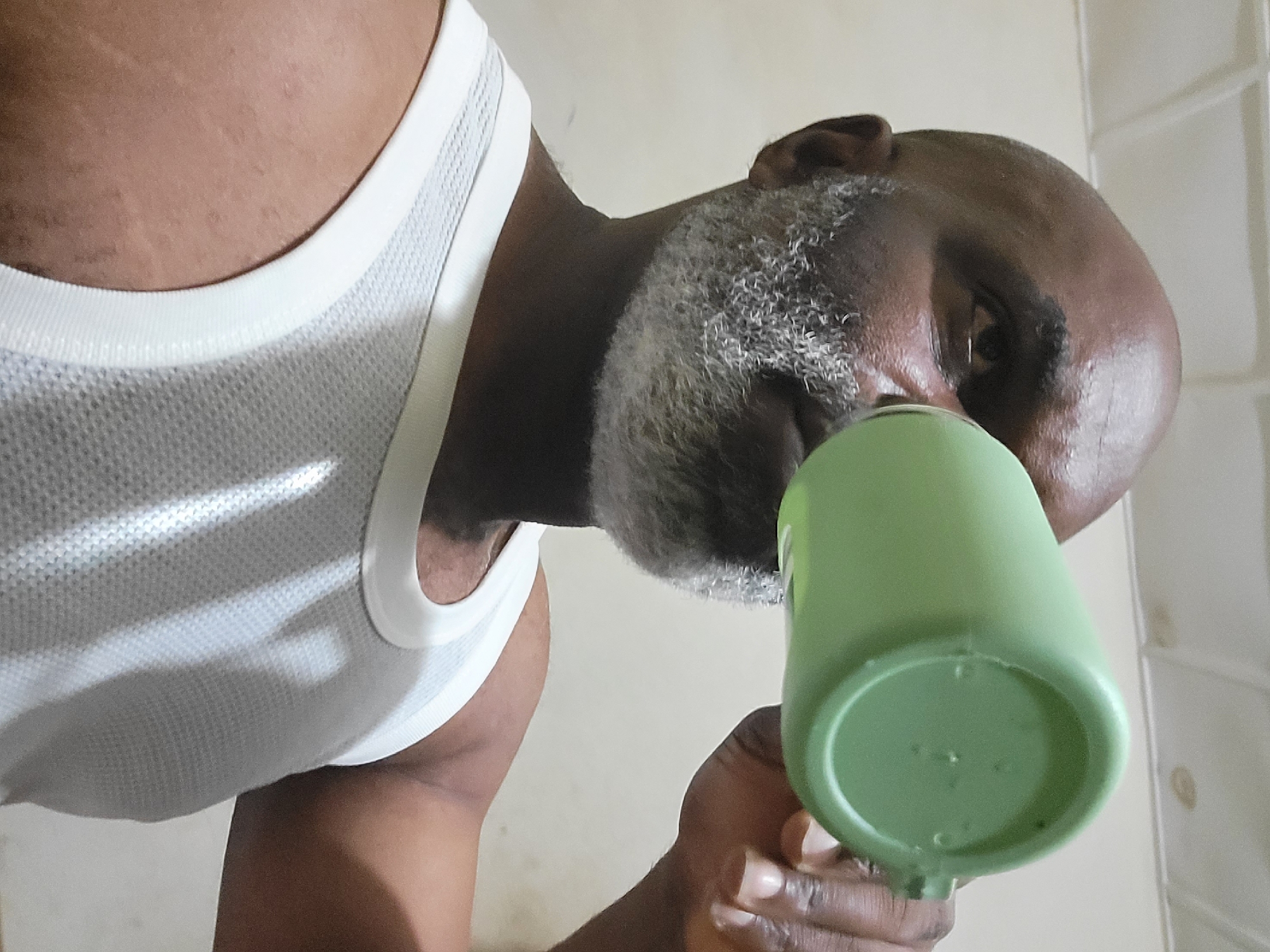 A low-angle close-up shows a Black man with a graying beard and a bald head drinking from a light green plastic cup. He is wearing a white tank top. The background consists of a beige wall. The location is Unknown, Unknown.