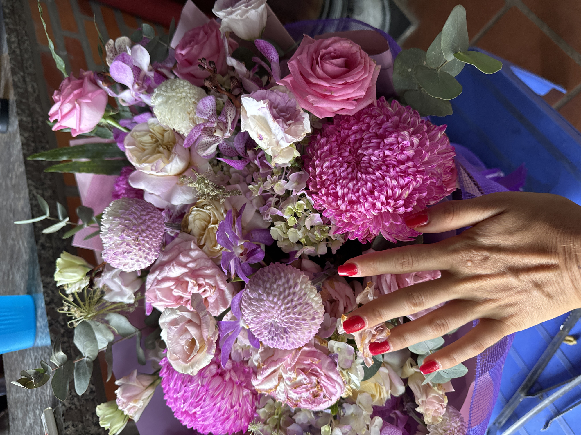 In Mũi Né, Vietnam, a close-up shot captures a vibrant bouquet of pink and purple flowers, with a woman's hand, adorned with bright red nail polish, resting on the arrangement. The hand is positioned to the left of the frame, obscuring some of the flowers. The bouquet features a variety of textures and forms, including fluffy chrysanthemums, delicate roses, and orchid-like flowers, interspersed with greenery. The background is blurred, but suggests an indoor setting with hints of a countertop, a blue object, and a brick wall. The overall impression is one of natural beauty and a touch of personal adornment.