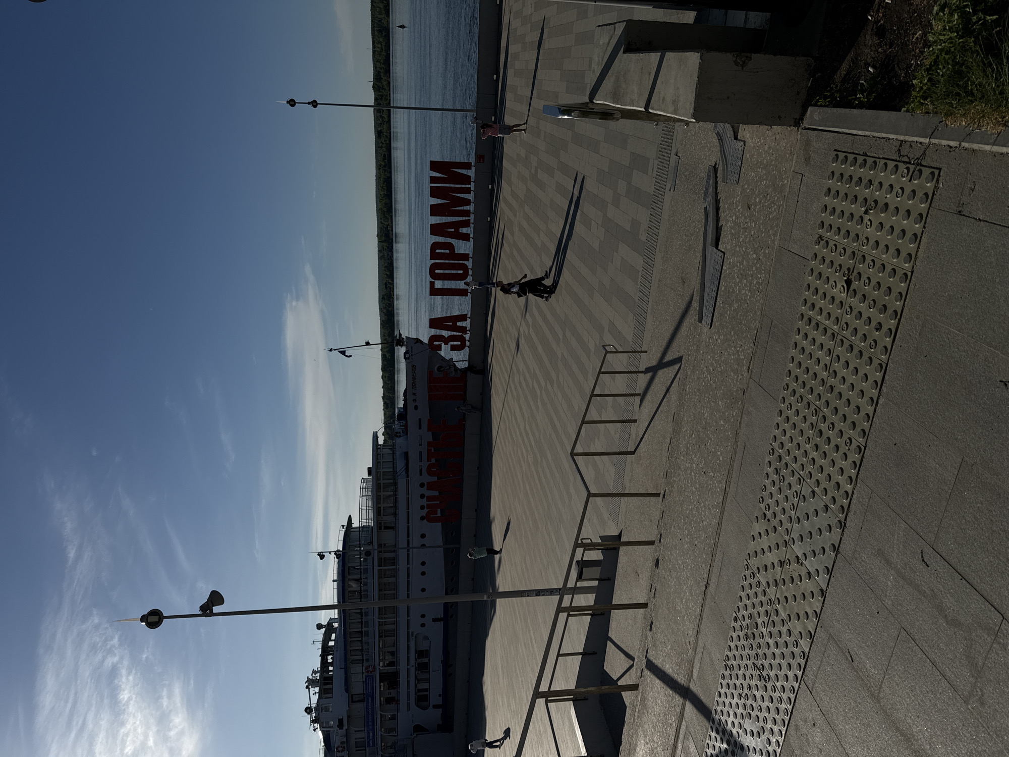 The image captures a waterfront scene in Perm, Russia. A large passenger ship is docked along the embankment, with the prominent Cyrillic inscription "ЗА ГОРАМИ" (ZA GORAMI) in red lettering displayed prominently on the embankment. Several people are walking along the paved embankment area, casting long shadows due to the angle of the sun. Street lamps line the embankment, and a body of water extends into the background. The paving consists of grey stone tiles and metal plates with tactile dot patterns. Handrails are visible along the side of the embankment.