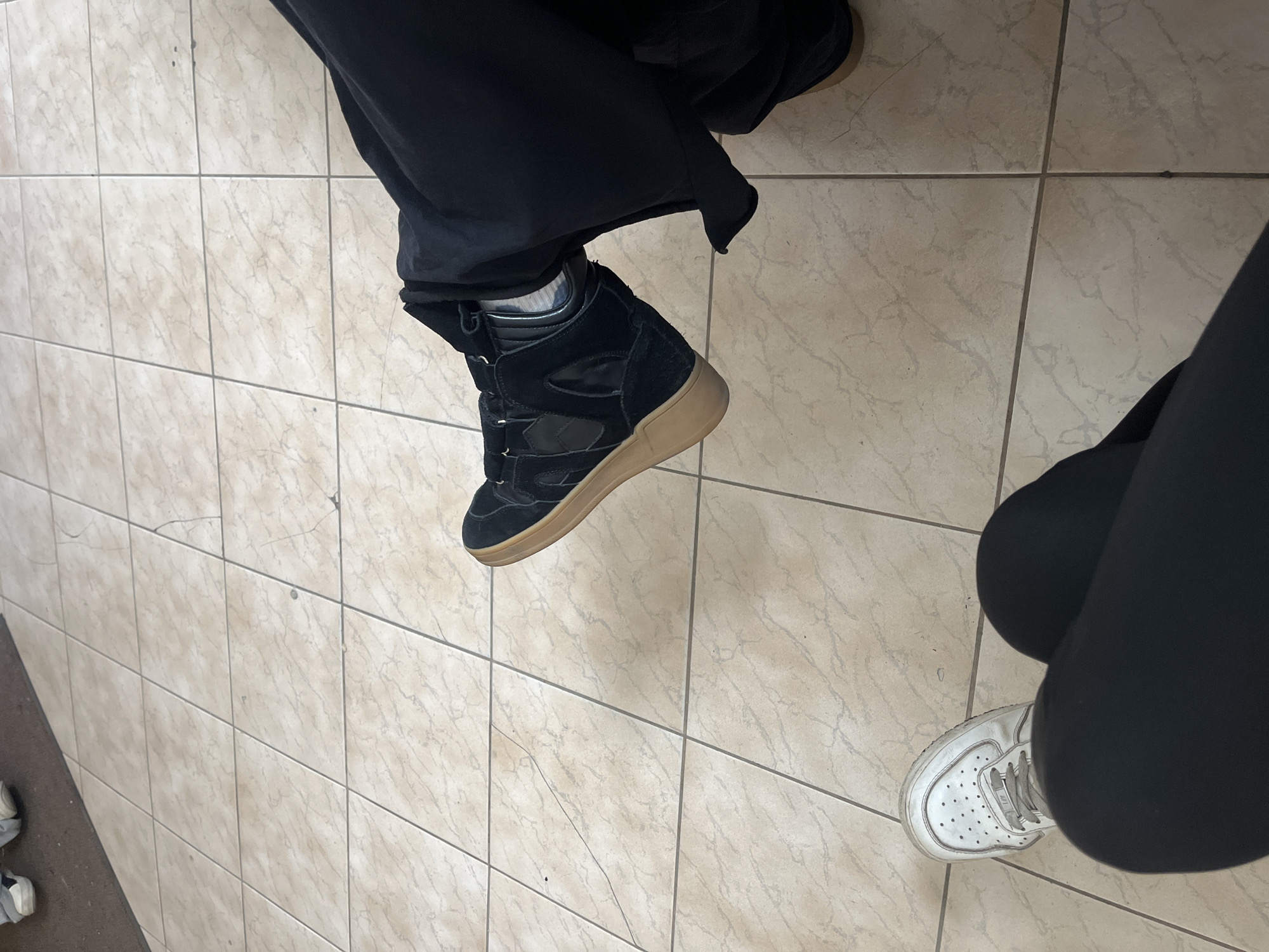 A top-down shot depicts two individuals indoors. One person wears black sweatpants and a black suede high-top sneaker with a tan sole. The other person is clad in black leggings and a white sneaker. The background consists of beige square floor tiles. The location is unknown and there is no evidence to suggest that it is in Saint Petersburg, Russia.