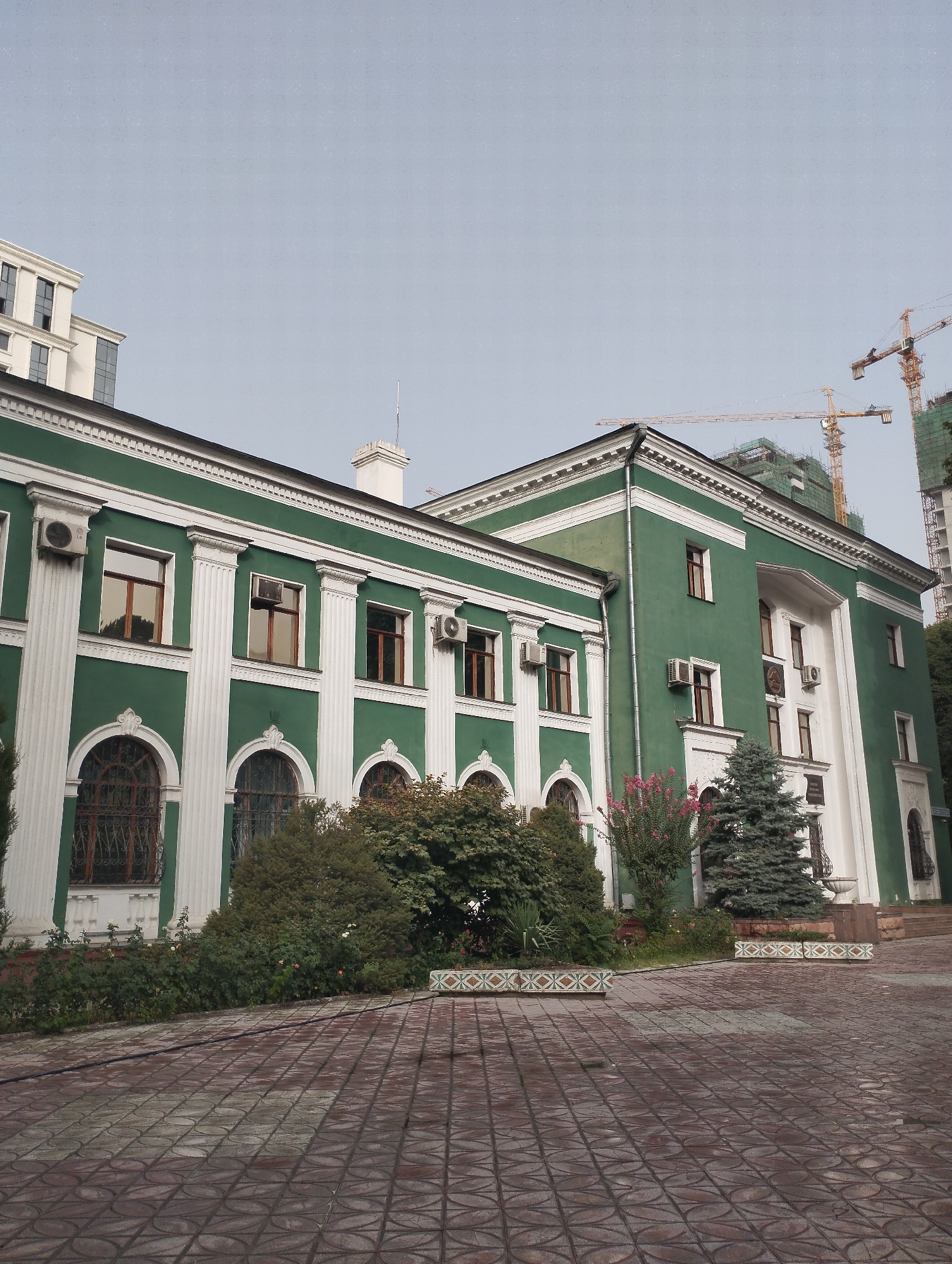 The image depicts the exterior of a green building with white decorative trim and columns. The building has several windows, some with arched tops and decorative grates. Multiple air conditioning units are mounted on the exterior walls. In the foreground, a paved courtyard with a patterned surface leads towards the building. Lush green bushes and trees, including a coniferous tree, are planted along the front of the building. In the background, two yellow construction cranes are visible against a pale sky, suggesting ongoing development or construction in the vicinity. A taller, modern white building is also partially visible on the left side of the image. A sign with text is mounted on the right side of the building, but the text is not legible. The scene appears to be a public space or institution.
This building is the Palace of Nations in Dushanbe, Tajikistan, which serves as the presidential palace. The construction cranes in the background indicate nearby development projects.