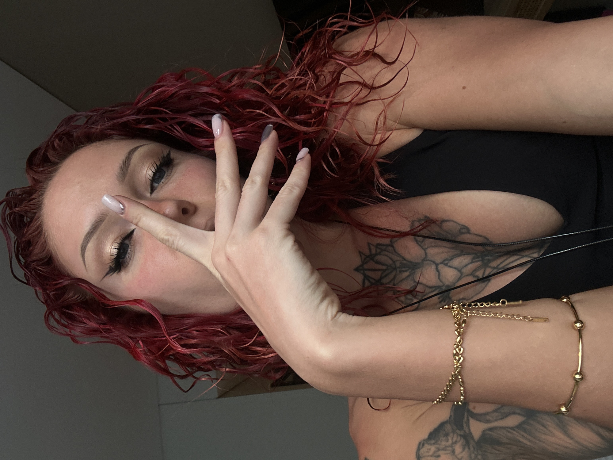 A red-haired woman with fair skin is indoors. She is wearing a black tank top and gold bracelets on her left wrist. Her left hand is held up in front of her face, with her fingers spread. She is wearing makeup, including eyeliner. The background consists of a plain wall. Her upper chest area shows tattoos. The location and city are unknown.