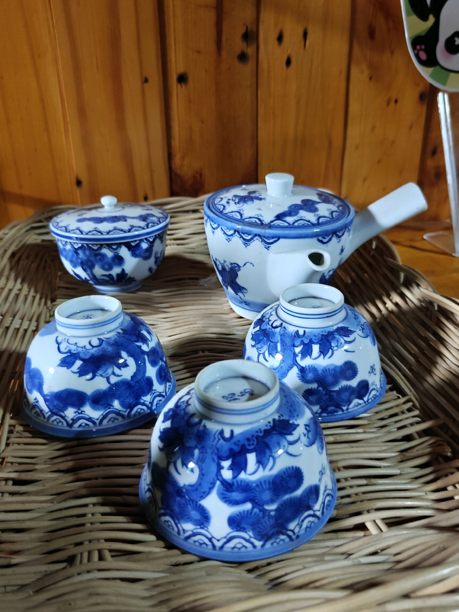 The image depicts a collection of blue and white ceramic ware, including a teapot with a handle and spout, and four bowls, arranged on a woven mat. The ceramics are decorated with traditional Asian motifs, featuring stylized plants and waves. The background consists of a wooden wall. The lighting is warm and natural, casting shadows. The scene is static and appears to be a still life arrangement, possibly for sale or display. There are no people or animals present. The location is not identifiable as Phra Nakhon Si Ayutthaya City Municipality, Thailand, based solely on the visual information provided in the image.