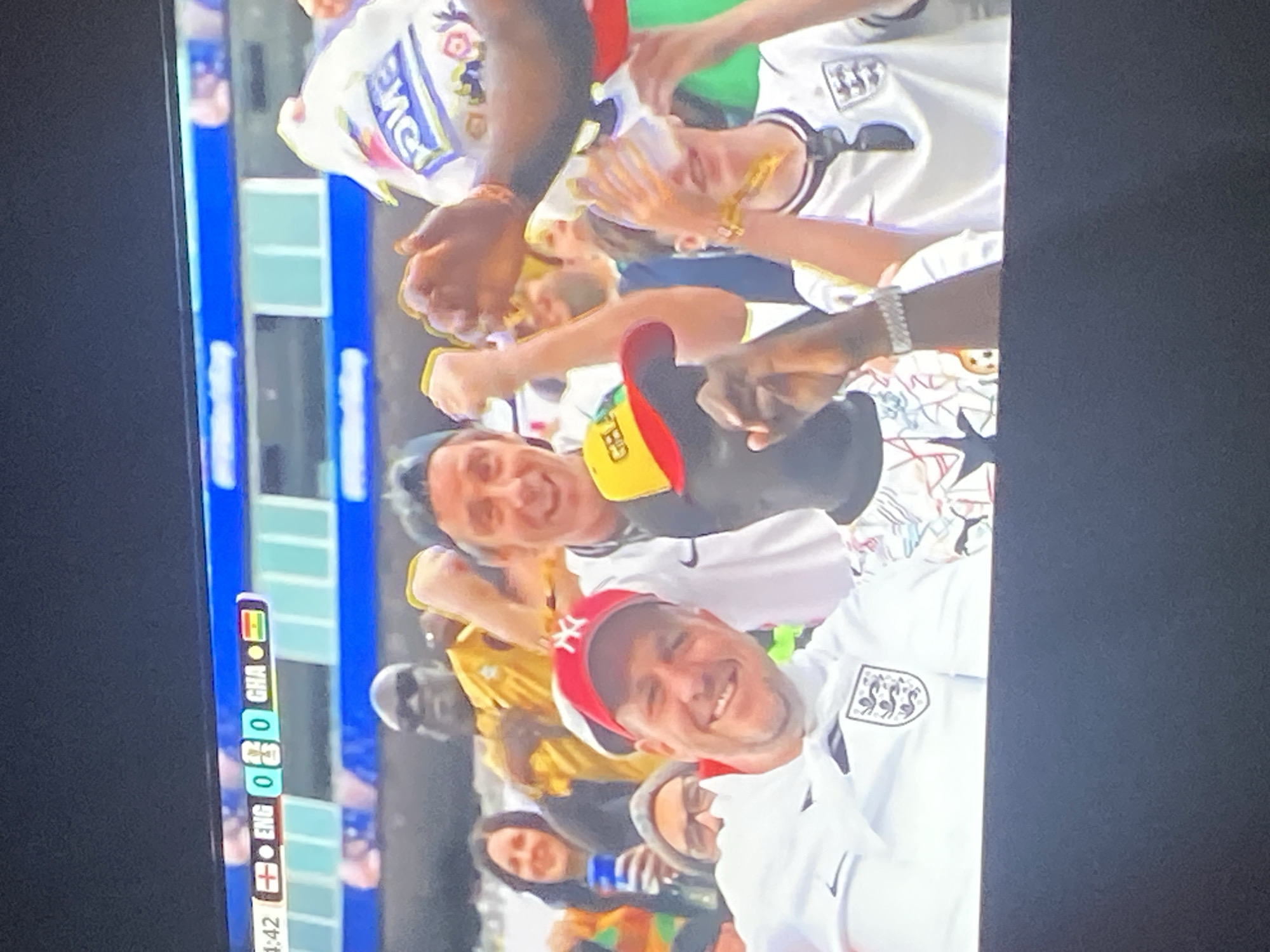 This media file shows a group of people in a stadium setting, likely at a sporting event given the scoreboard visible on the left. The scoreboard displays "ENG 0" and "GHA 0," indicating a match between England and Ghana with a score of 0-0. The people appear to be fans, some wearing England jerseys with the team's crest. One individual in the foreground is wearing a red and white baseball cap and smiling, while others around them also display happy and excited expressions. The overall atmosphere seems celebratory. The lighting suggests it is daytime. The visible text on the scoreboard is "ENG," "GHA," and "0" along with the flags of England and Ghana. The location context provided is Kuson, Nigeria, which aligns with the presence of Ghanaian fans.