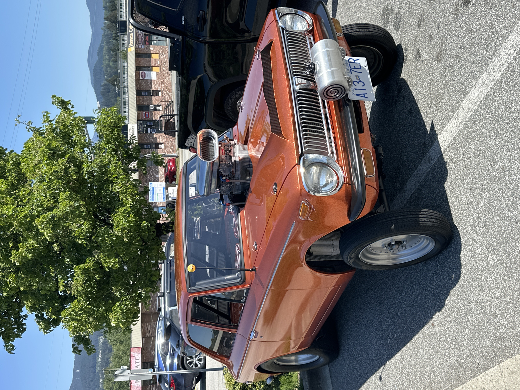 The image shows an orange modified vehicle in Port Moody, British Columbia, Canada. The car has an exposed engine and a British Columbia license plate, "A13 7ER". It's parked on pavement near a tree, other parked cars, and a multi-story brick building with commercial signage visible. Mountains are in the background.