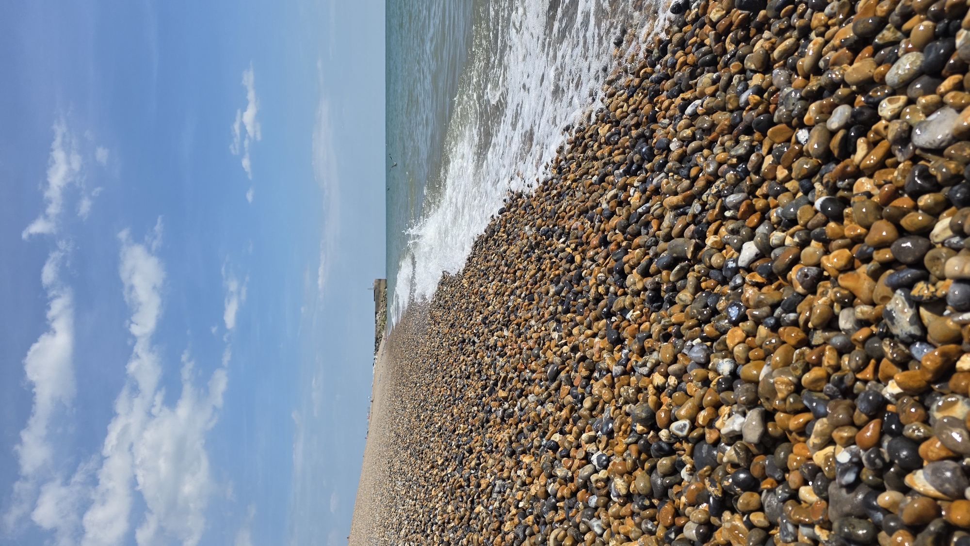 A wide, low-angle shot captures a dense, colorful pebble beach stretching into the distance in Hastings, United Kingdom. The pebbles, ranging in color from light tan to dark grey and black, are wet and glistening from the gentle waves of the sea. The sea itself is a deep teal, with a white foamy wave breaking on the shore, creating a dynamic line between the land and water. In the background, a concrete pier or sea defense extends out into the water, with a small structure at its end. The sky above is a clear, bright blue, dotted with fluffy white clouds. A few birds are visible flying above the water. The overall impression is one of a sunny, breezy day at the coast.