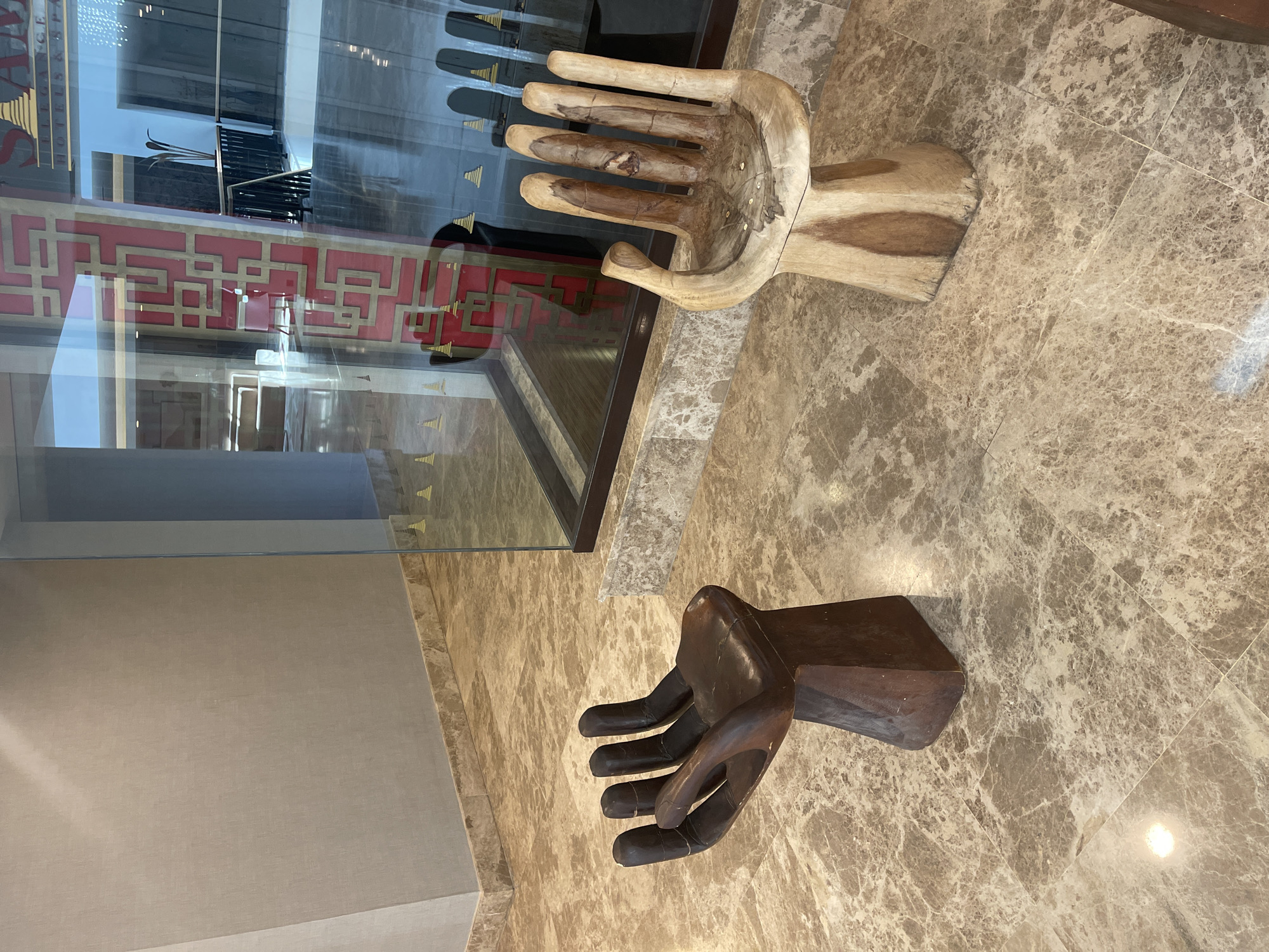 The image shows two wooden hand sculptures on a marble floor in an interior setting. One hand is light-colored and open, while the other is dark and closed. A glass display case with a red and gold design is visible behind the light-colored hand. The exact location in the city of Unknown, Unknown is not discernible from the image.