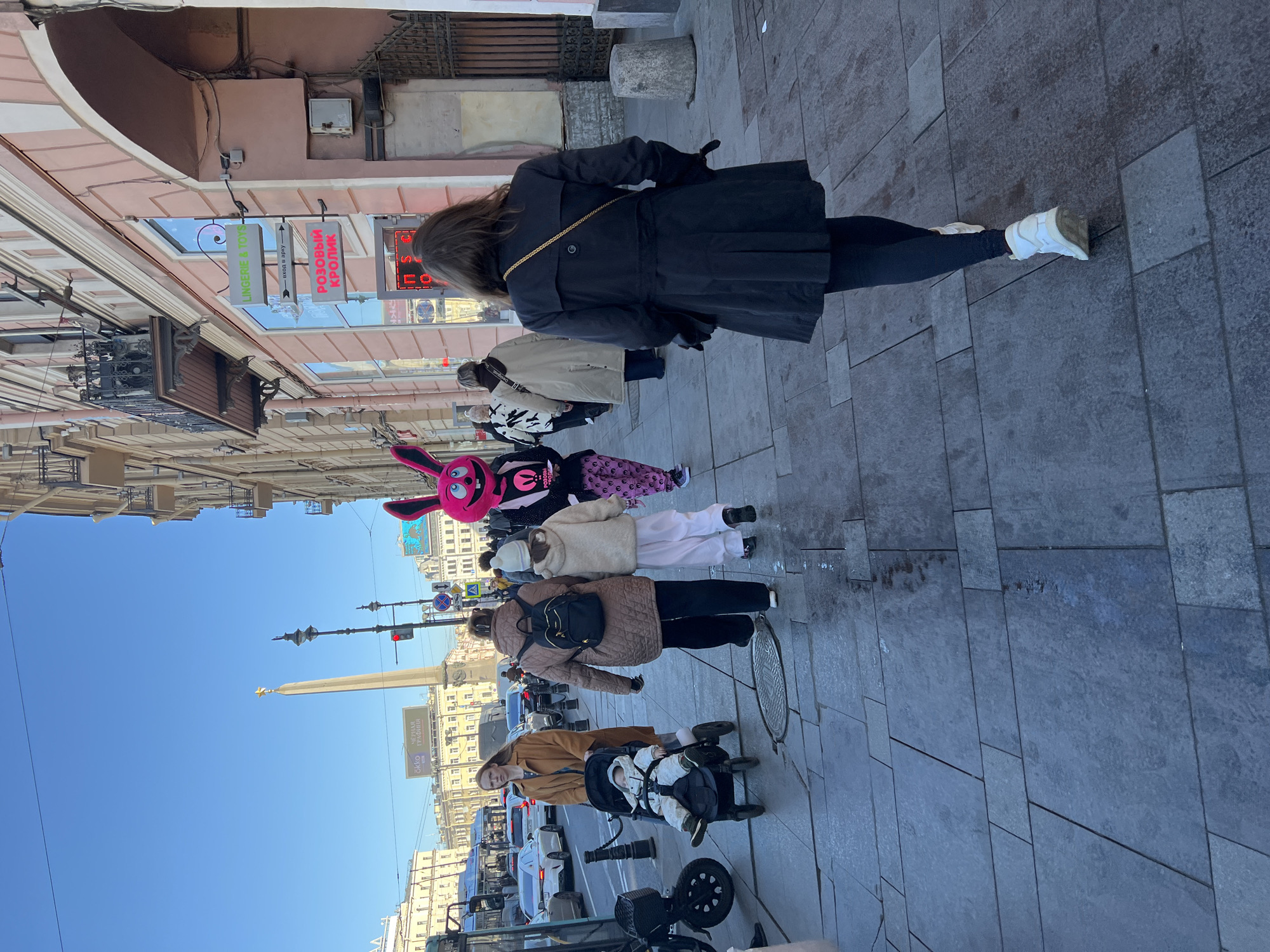 The image captures a street scene in Saint Petersburg, Russia, showcasing several individuals walking along a tiled sidewalk on a clear, sunny day. The foreground features a woman walking away, wearing a black trench coat, dark leggings, and white sneakers. A gold chain is visible across her back. In front of her, several other people are walking. One of them is wearing a pink bunny head. There is a woman pushing a baby in a stroller. In the background, an obelisk stands prominently against the blue sky. Buildings line the street, with signs in Russian visible, including a shop sign that says "РОЗОВЫЙ КРОЛИК" or "PINK RABBIT" in English. Parked vehicles are visible along the street edge, indicating traffic in the area.