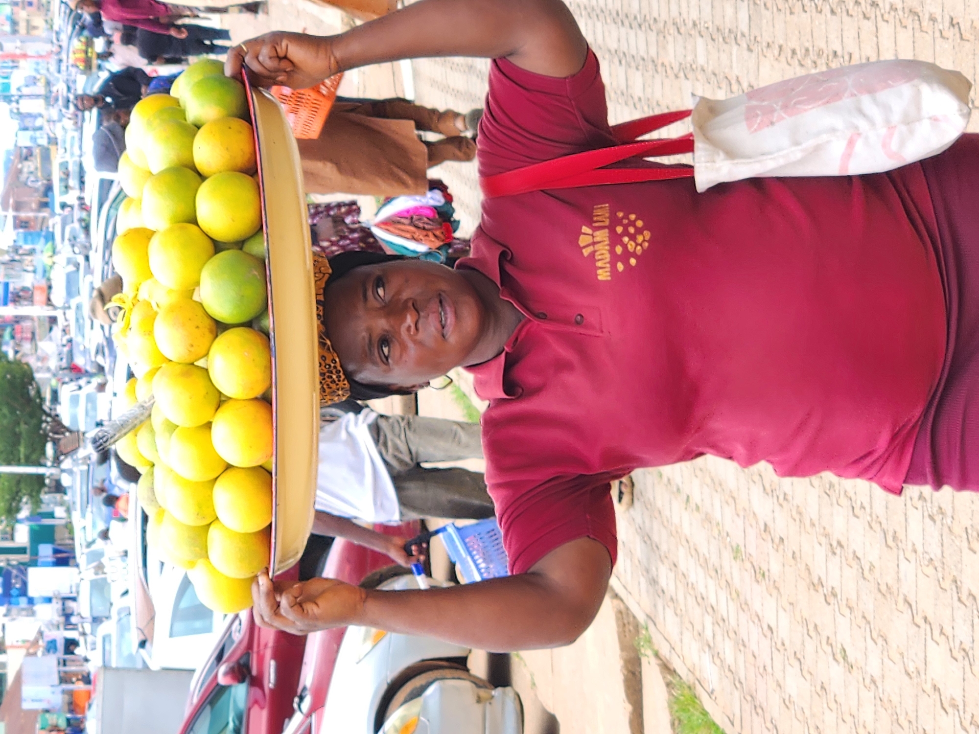 A woman is pictured outdoors in a city, carrying a large tray of bright yellow and green fruits on her head. She is wearing a maroon polo shirt with a yellow logo that reads "MADAM LAMI" and a small yellow and brown patterned headscarf. A white tote bag with pink markings hangs from her right shoulder, its strap a vibrant red. Behind her, the scene depicts a busy street with multiple vehicles, including cars and possibly a truck, along with other pedestrians in the background. The ground is paved with interlocking concrete blocks. The environment suggests a market or a bustling urban area.