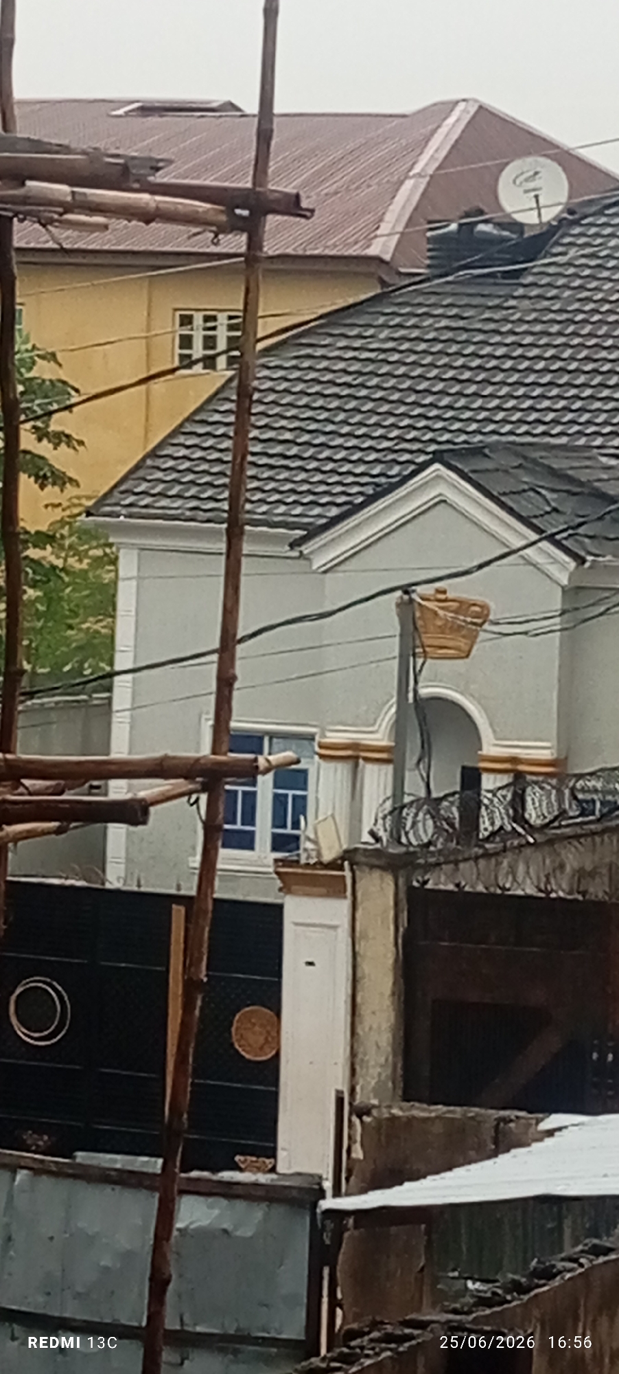 The image shows an outdoor scene with several houses. In the foreground, there is scaffolding made of bamboo poles. Behind the scaffolding, a light gray house with a dark gray tiled roof is visible. This house has a white decorative trim around the eaves and a window with blue glass. A black metal gate with a circular design is in front of the house, and there's a round, woven decorative object on the gate. Next to the gray house, a yellow house with a brown roof and white-framed windows can be seen. In the background, another brown roof is visible, with a satellite dish and a water tank on it. The weather appears overcast, suggesting it might be a cloudy day or early morning/late evening. The image also contains timestamps and text at the bottom: "REDMI 13C" and "25/06/2026 16:56". The overall impression is of a residential area with ongoing construction or renovation work. There are no people visible in the image.