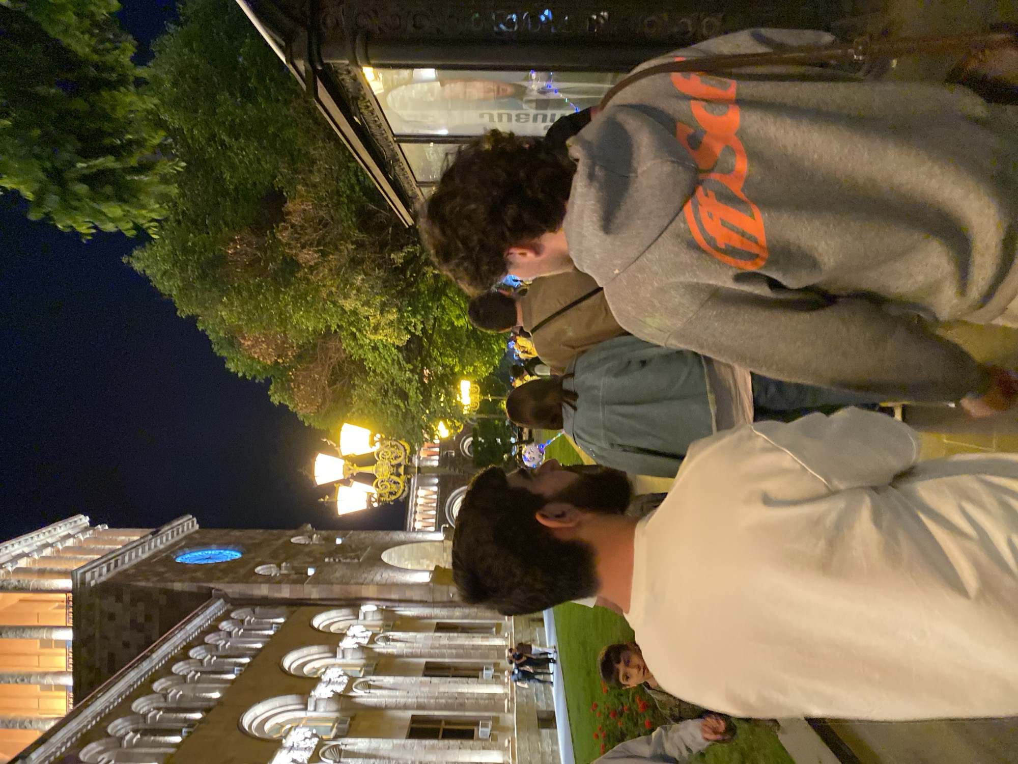 The image depicts an outdoor evening scene in Yerevan, Armenia, illuminated by artificial lights against a dark night sky. Several people are present, with two individuals prominently featured in the foreground, seen from behind or in profile.
On the left, a person with dark, curly hair wears a grey hooded sweatshirt with a bold orange graphic on the back, where parts of the letters "GE" or "ES" are discernible, likely forming part of a word like "ORANGE" or "ESSAGE". This individual appears to be looking towards the right side of the frame. Behind them, a storefront window reflects parts of the street and shows reversed text, possibly "anenu" or similar characters in a non-Latin script, suggesting it could be Armenian.
To the right and slightly closer to the viewer, another person with dark hair and a beard is seen in profile, wearing a plain white long-sleeved shirt. In the lower right corner, a child's face is partially visible, looking towards the camera. Other blurred figures are scattered in the mid-ground, indicating a public space with a number of people.
The background is dominated by a large, ornate building with classical architectural features, including arches, columns, and decorative cornices. The building is warmly lit, highlighting its details. A distinctive round window with a blue light or glass is visible on an upper section of the building. Ornate, golden street lamps with multiple bulbs provide further illumination to the scene and the surrounding area. To the left, dark green trees stand against the clear night sky. The scene suggests a pleasant evening atmosphere in an urban environment.