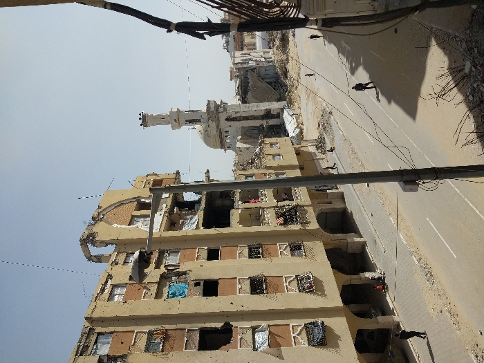An elevated view captures a street scene in Khan Yunis, Palestinian Territory, marked by extensive structural damage under a clear daytime sky. In the foreground, a utility pole stands on the right, secured with wires. The ground directly beneath shows scattered debris, including exposed rebar and concrete fragments.
On the left, a multi-story building with a light beige facade exhibits severe damage across its visible five to six floors. Numerous perforations and impact marks mar its exterior walls. Many windows are shattered or missing entirely, with some frames bent or dislodged. Portions of the upper structure are collapsed, revealing interior rooms, and an arched architectural element at the roofline is partially destroyed. Some windows and balconies display hanging fabrics or makeshift coverings. Below the building, an arcade formed by ground-floor arches shelters a small group of two to three individuals, possibly children, seated on the sidewalk.
A paved road with a central white double-line marking stretches through the midground, appearing dusty with sand. Several individuals are dispersed across the scene: one person walks on the sidewalk in the lower-left, another strides in the center of the road, and two more are further down the street on the right. Additional figures are visible on the right-side sidewalk.
In the background, behind the damaged building on the left, a prominent white mosque stands, featuring a large dome and a tall, slender minaret. The mosque also shows signs of damage, particularly around its upper sections. Beyond the mosque, the skyline reveals further urban structures, some appearing damaged, under the bright sky.
