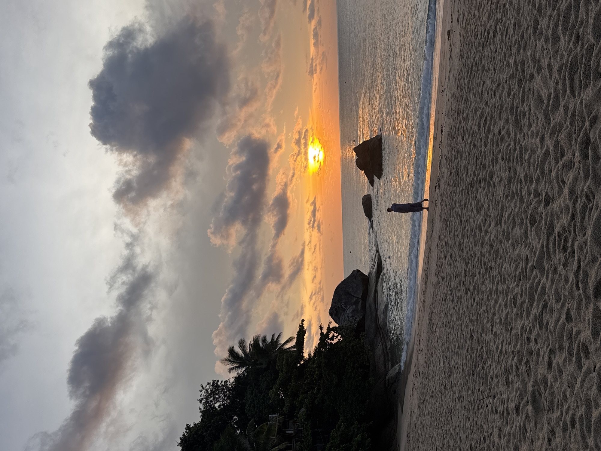 The image shows a beach scene at sunset in Baan Thong Nang, Thailand. The sun is partially obscured by clouds, casting an orange glow on the sky and water. Several large rocks are visible in the water near the shoreline. A person stands on the sand near the water's edge, facing the ocean. The beach is sandy with wave patterns. Trees and other vegetation line the edge of the beach. The overall lighting is dim due to the sunset and clouds.