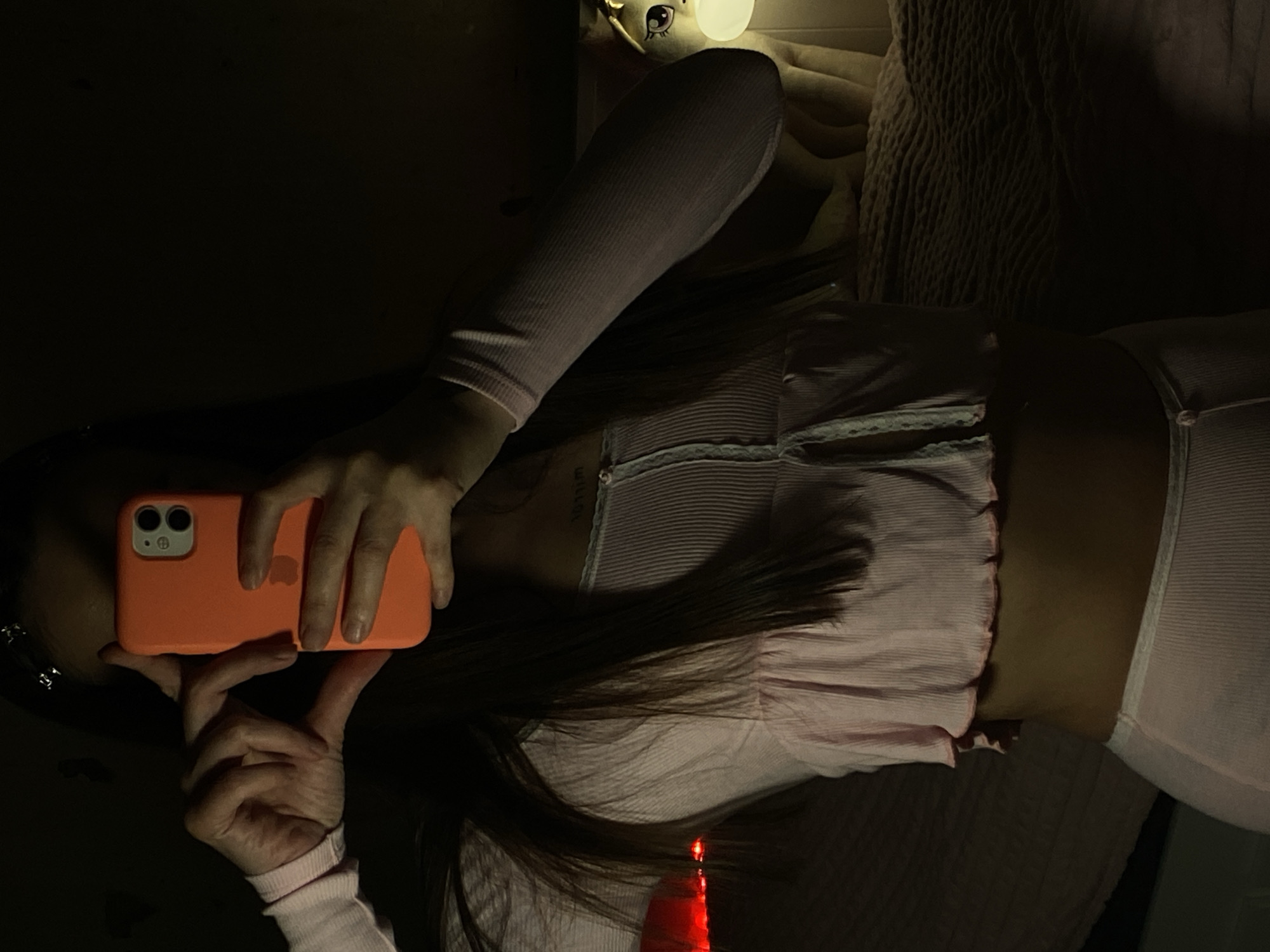 A woman is lying on her side in a dimly lit room, holding an orange mobile phone in front of her face, presumably taking a selfie. She is wearing a pink, long-sleeved top and matching bottoms. Some of her long, dark hair is visible around the phone and over her top. A small red object, possibly a candle, is near her head. Behind her are what appear to be a plush toy and a textured blanket or pillow. The location is unknown, and there is no information in the image to suggest it is in Votkinsk, Russia.