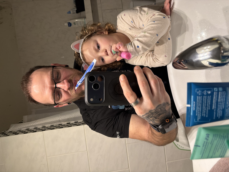 This image captures an adult male and a young child brushing their teeth, reflected in a bathroom mirror above a sink, located in Trassem, Germany.
The adult male, positioned on the left, has a beard, wears black-rimmed glasses, and has short dark hair. He exhibits ear gauges and tattoos on his left forearm. He is dressed in a black polo shirt. His left hand holds a black smartphone, featuring multiple camera lenses and a black protective case, oriented towards the mirror as if taking a selfie. A silver-toned ring is present on his left ring finger, and a smartwatch with an orange button is on his left wrist. He holds a blue and white toothbrush in his mouth.
The young child, situated to the right of the male, has curly brown hair and wears a light-colored headband adorned with two light pink, glitter-accented cat ears. She is dressed in a light-colored long-sleeved shirt patterned with small, multi-colored hearts. She holds a pink and green toothbrush in her mouth. Both individuals are actively brushing their teeth.
The bathroom environment features white tiled walls. A shower curtain with a black and white checkered pattern is visible in the reflection behind the subjects. The white ceramic sink occupies the lower foreground, equipped with a chrome faucet. On the sink's surface, two tubes of toiletries, one green and one blue, are visible. In the background reflection, a white bottle and other containers are present on a shelf.