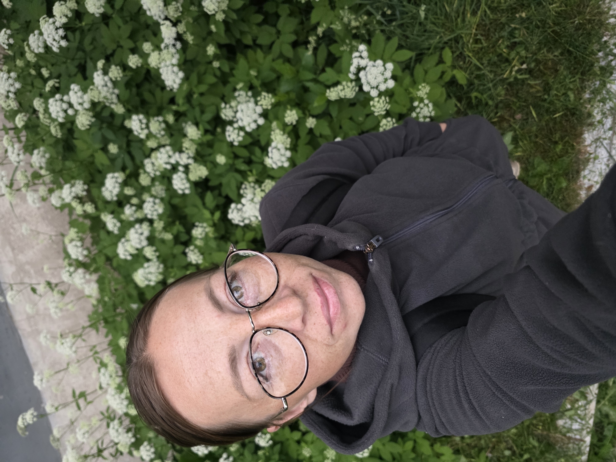 This image captures a high-angle selfie of an adult female standing outdoors within the city of Chaikovsky, Russia.
The woman is positioned slightly off-center, dominating the foreground and middle ground of the image. She has fair skin with a notable scattering of freckles across her forehead, nose, and cheeks. Her dark brown hair is pulled back, revealing her forehead and a central part. She wears round, wire-rimmed glasses with dark tortoise-shell accents and clear lenses, through which her light brown eyes are visible, directed upwards towards the camera. Her lips are closed in a relaxed expression, appearing to have a muted pink or nude color. She is dressed in a dark grey (charcoal) fleece hooded jacket with a front zipper, beneath which a brown turtleneck or high-collared garment is visible at her neck. A dark backpack is worn, with its straps visible over her shoulders, partially obscured by the jacket.
The background is filled with dense green foliage interspersed with abundant clusters of small white flowers, characteristic of a flowering herbaceous plant, possibly goutweed or a similar umbellifer. This plant growth extends upwards and to both sides of the woman. In the upper left background, behind the foliage, a blurred, light-colored wall or concrete surface is visible. The ground beneath the flowers and to the lower right consists of green grass. A light-colored concrete or stone edge is present along the bottom and lower left of the frame, separating the grassy area.